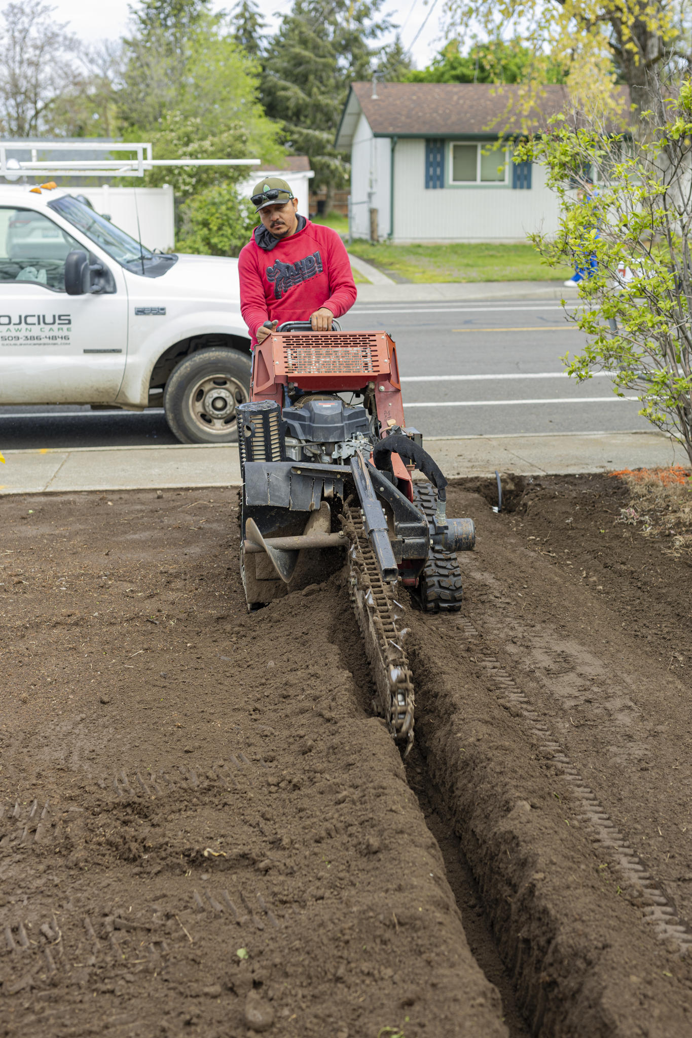 A man in a red hoodie and green cap operating a mini excavator on a construction site in front of a residential neighborhood.