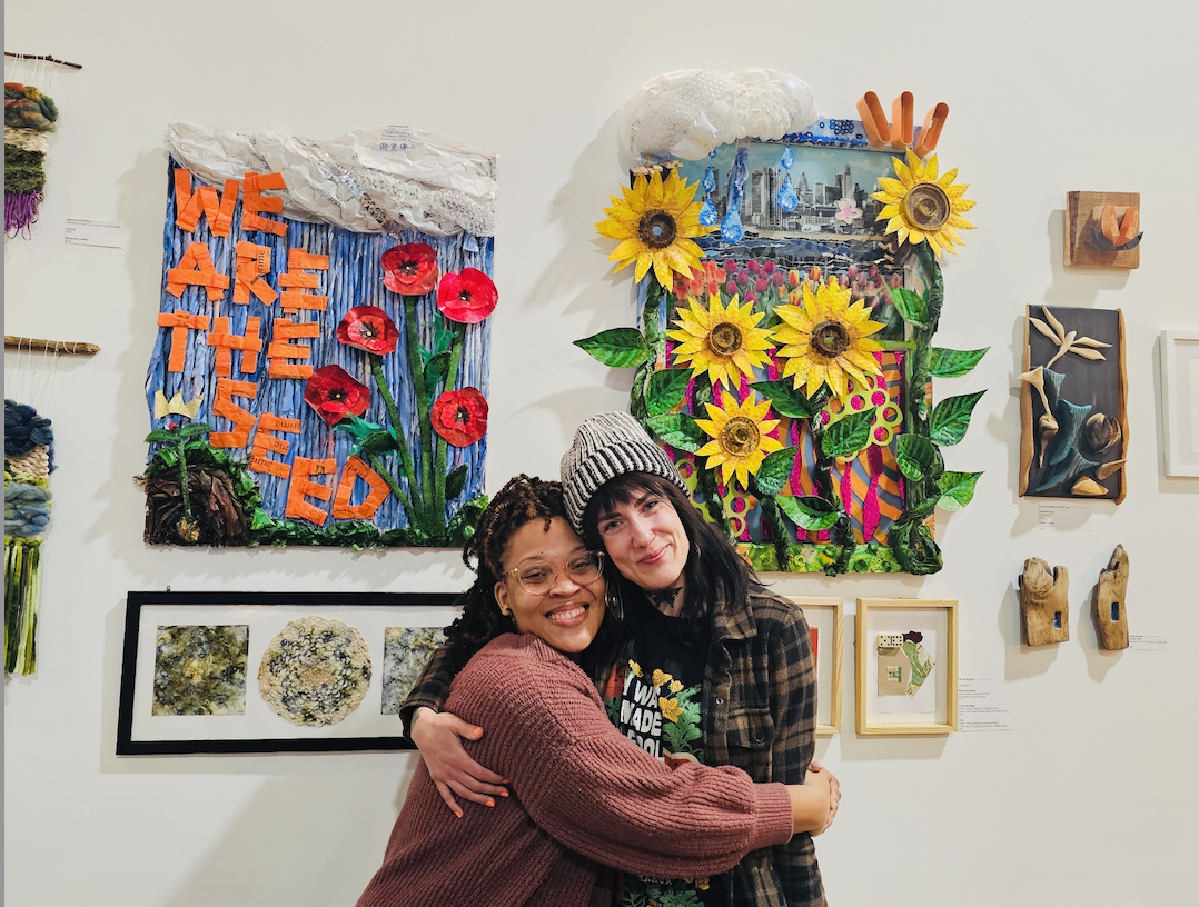 Two women hugging and smiling in front of colorful wall art at an art gallery, featuring flowers, text, and mixed media elements.