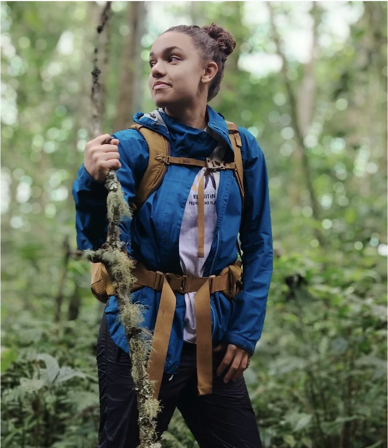 Young woman with curly hair in a bun exploring a dense green forest, holding a moss-covered branch, wearing a blue jacket and a brown backpack.