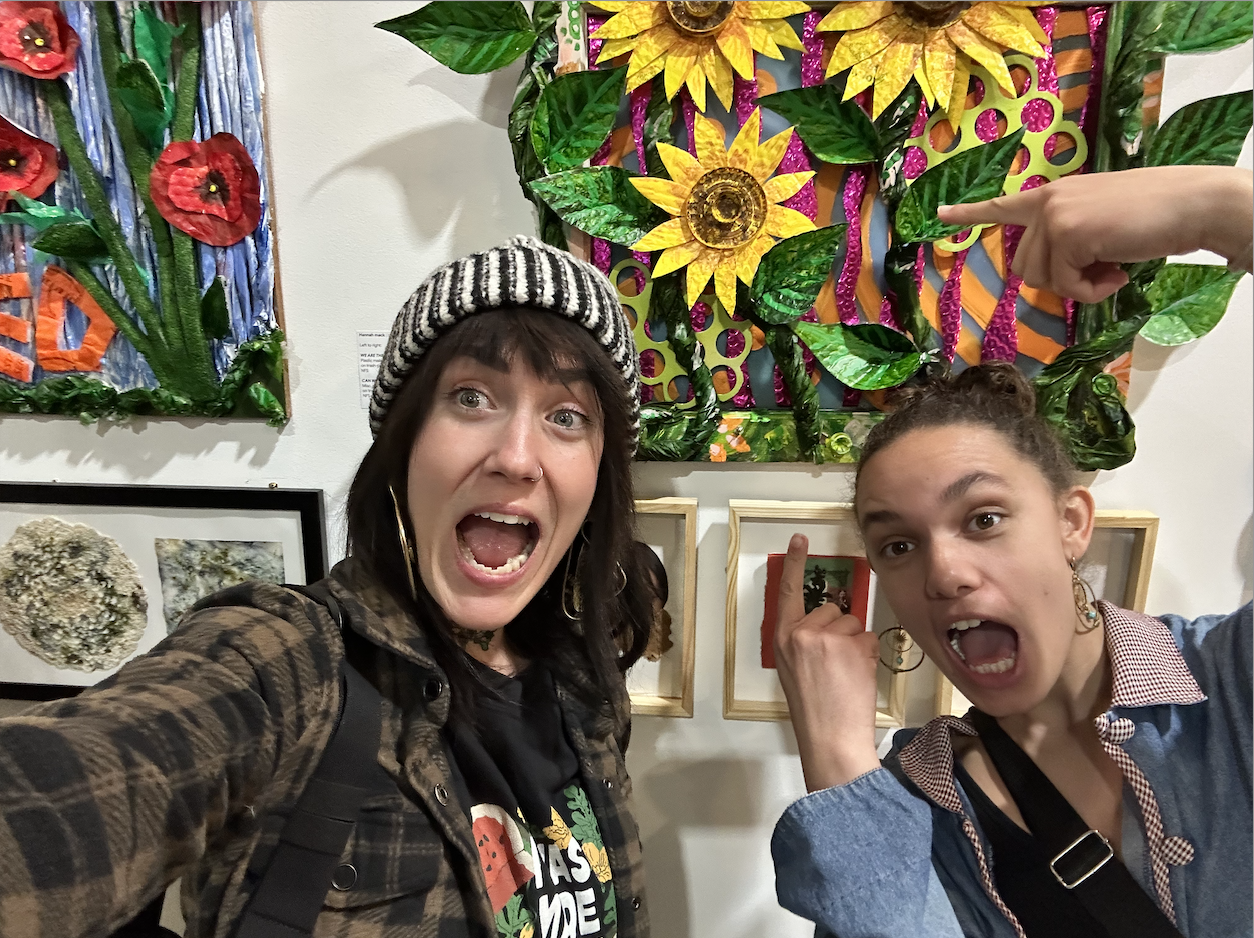 Two women taking a selfie at an art gallery, with colorful floral and nature-themed artwork on the wall behind them, both making expressive faces
