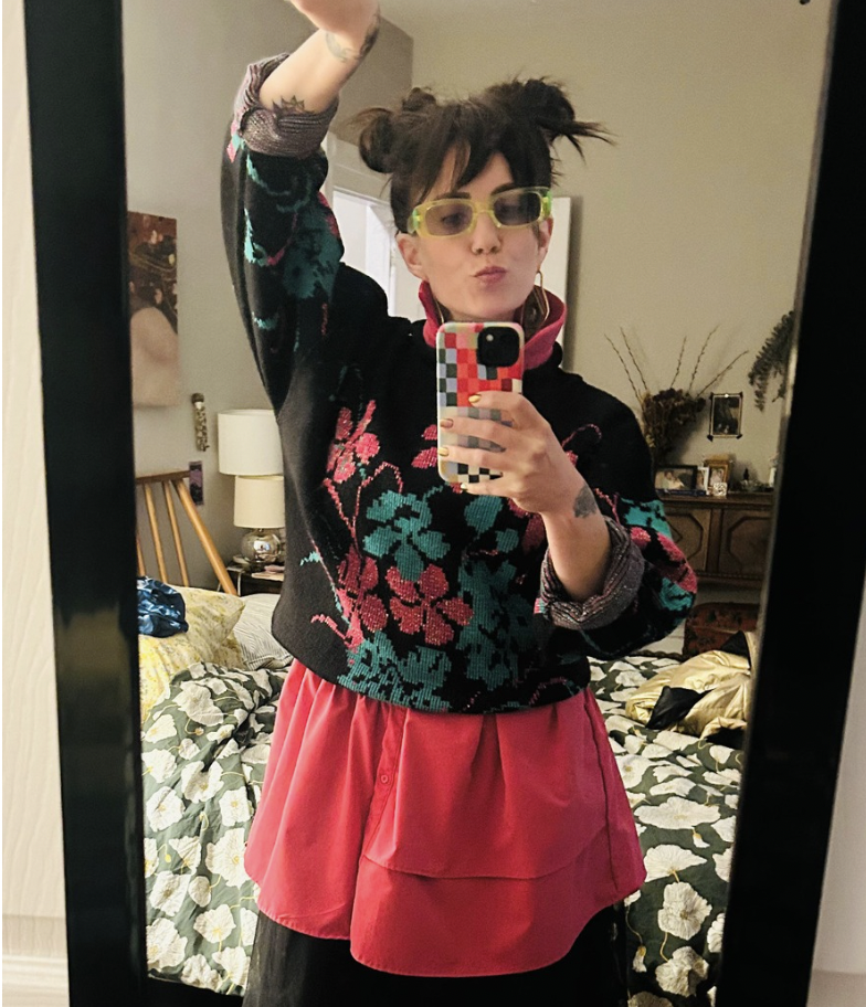 Person taking a mirror selfie in a bedroom. They have short, messy dark hair with two small buns, wear yellow glasses, a black sweater with a floral pattern, and a pink skirt. The background shows a bed with a floral bedspread and various bedroom furniture.