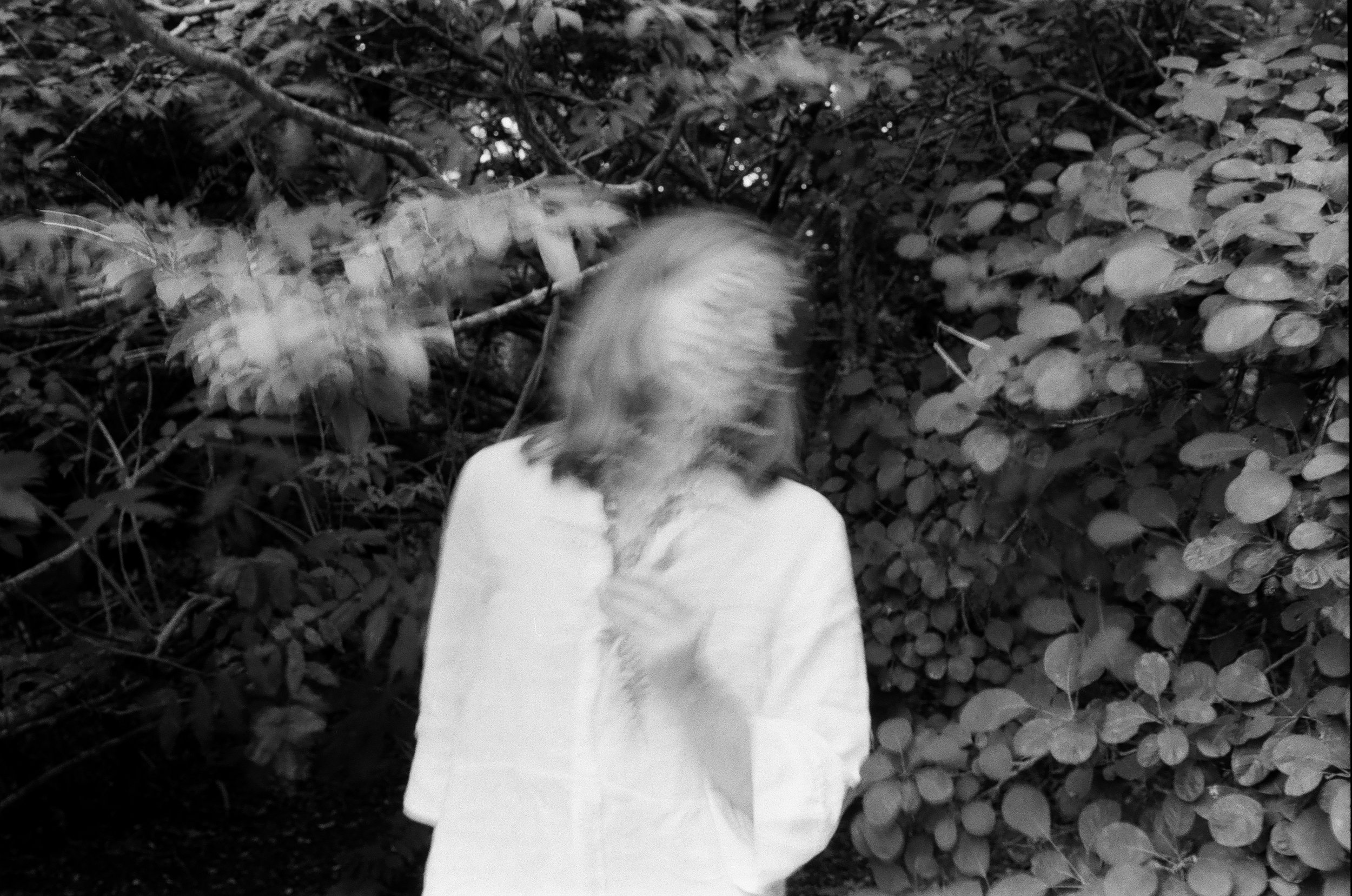 A person with shoulder-length hair in a white shirt standing in front of dense foliage, with their face obscured by their hair and hand.