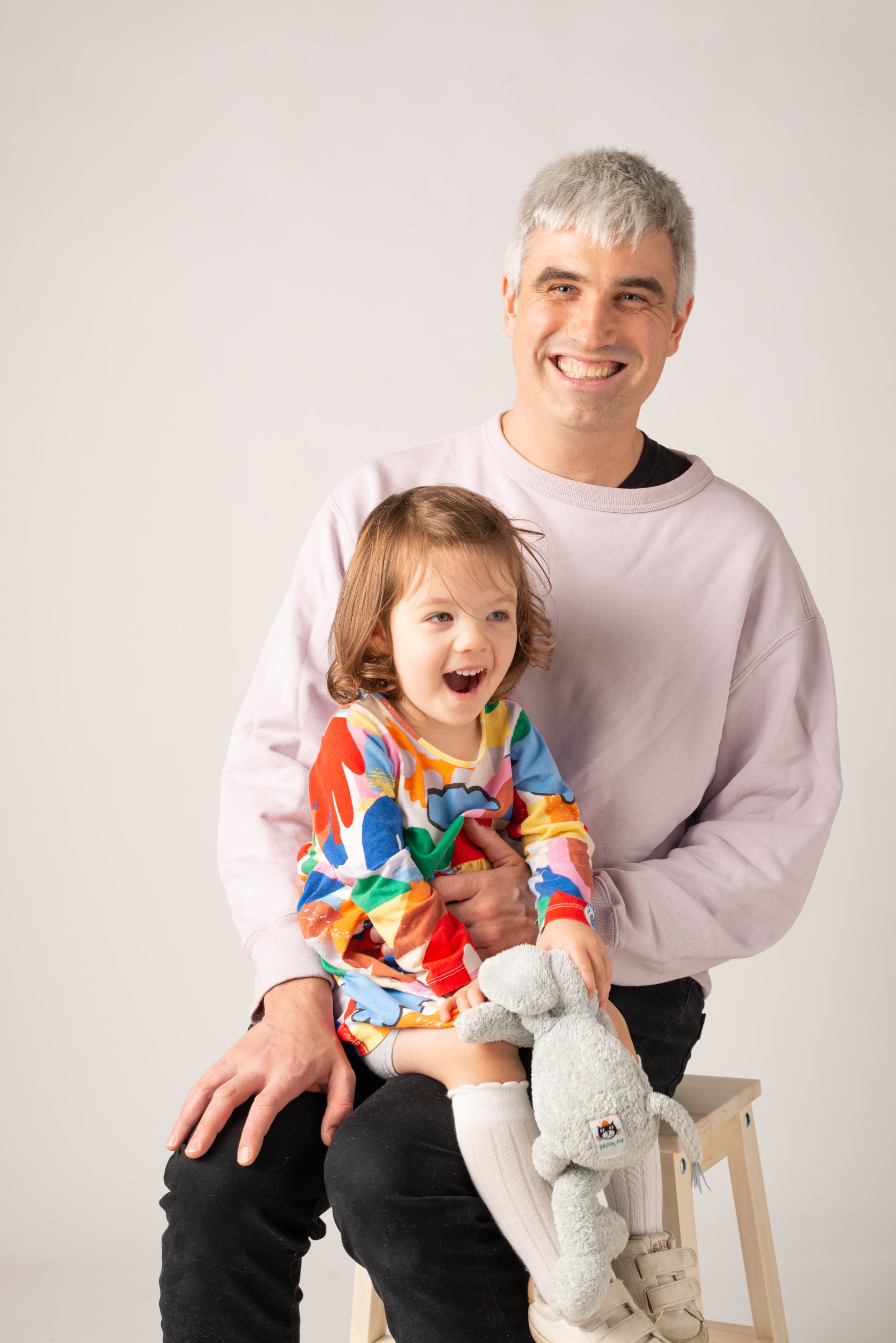 A man with gray hair and a young girl with brown hair sitting on his lap, both smiling and laughing. The girl is holding a gray stuffed elephant toy and wearing a colorful dress with white socks and white sneakers. They are sitting on a small wooden stool against a plain light background.