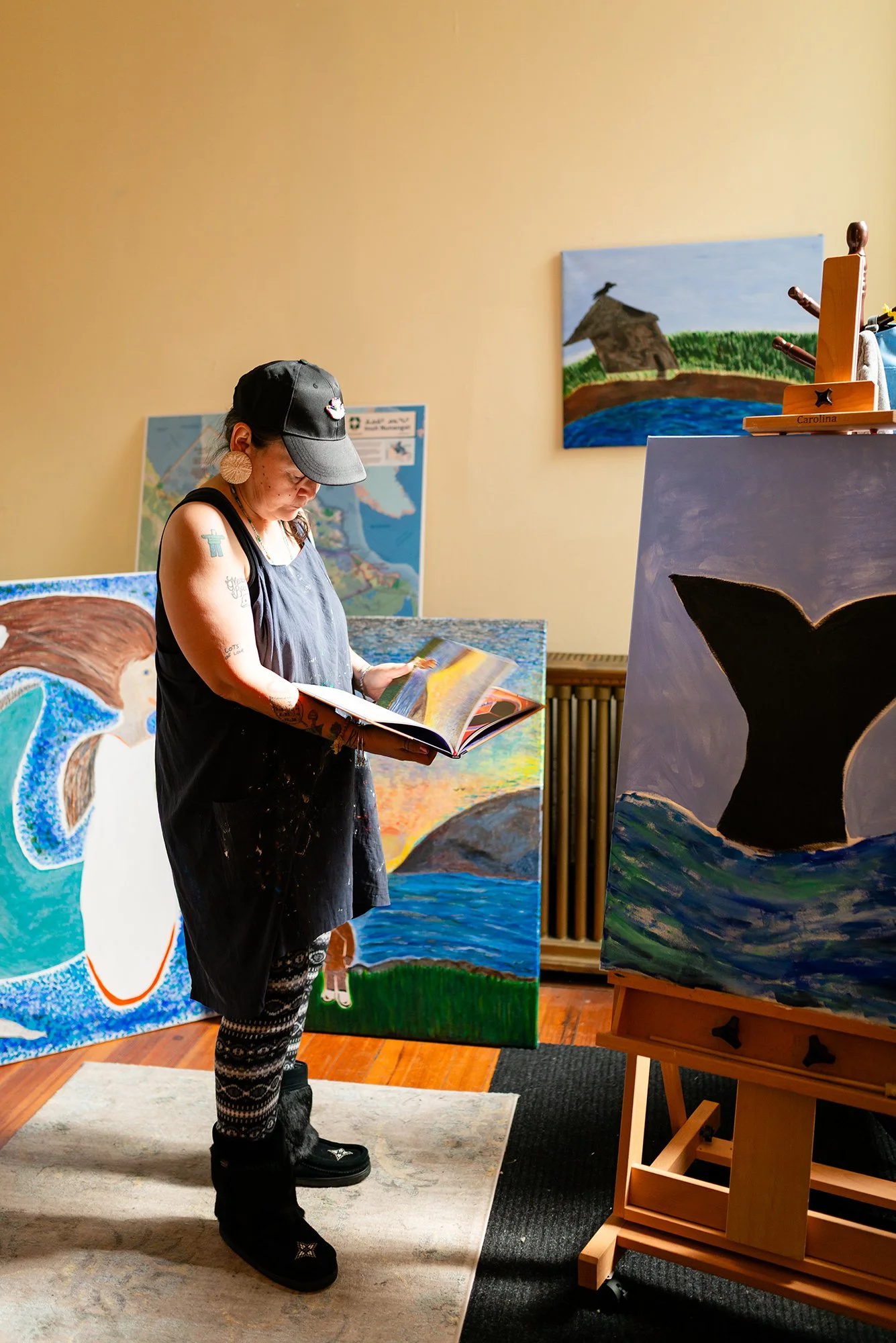 A woman wearing a black cap, earrings, and a black apron stands in an art studio reading a book. Behind her, several colorful paintings featuring seascapes, landscapes, and animals are displayed. To her right, an easel holds a painting of a whale, and a wooden art supply holder labeled "Carolina" is on a table.