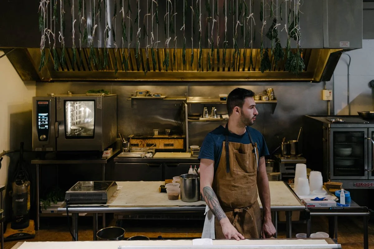 A man wearing a brown apron stands in a professional kitchen with various cooking equipment, including an oven, shelves, and containers with ingredients.