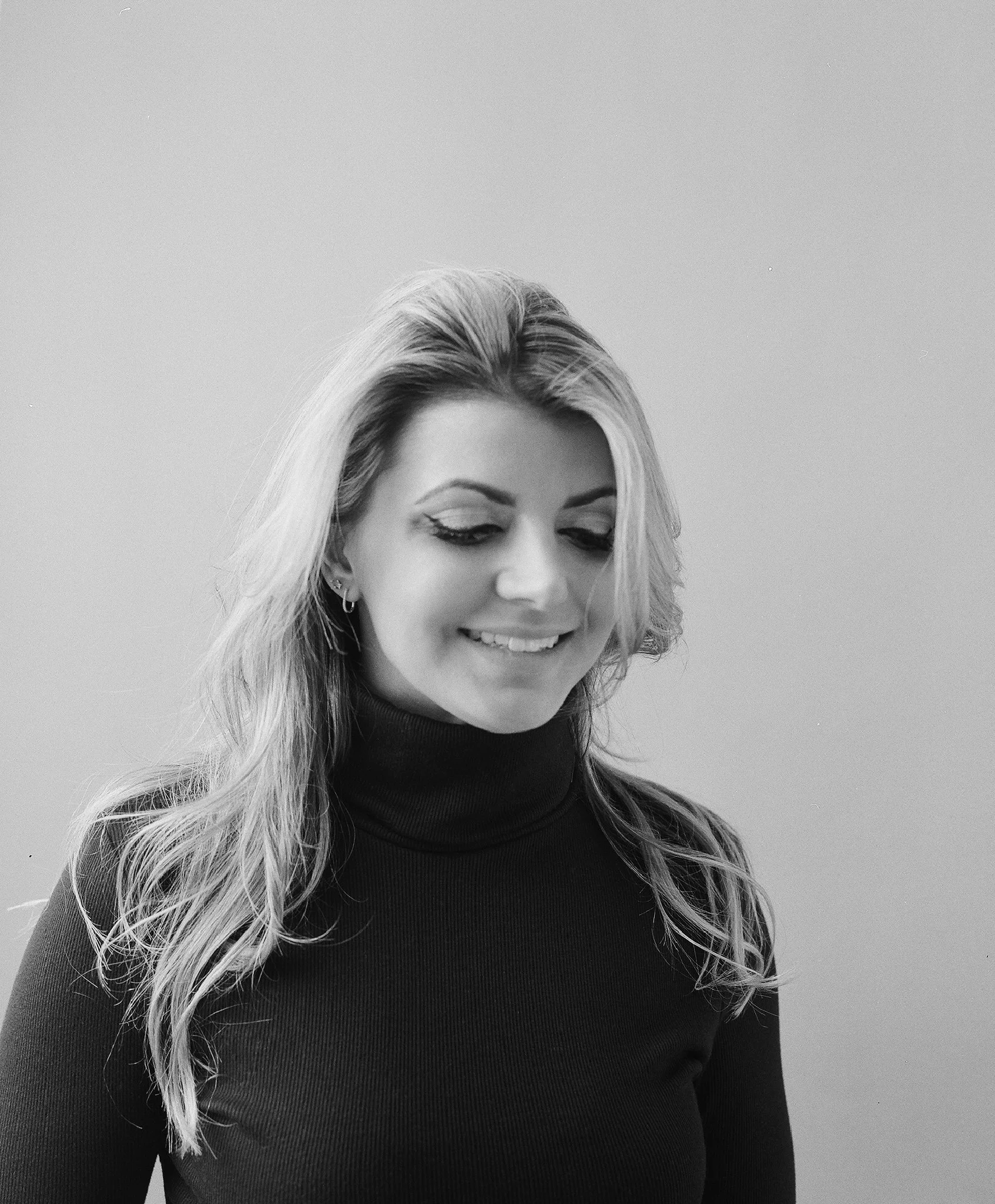 Black and white photo of a smiling woman with long hair wearing a turtleneck sweater, looking downward.