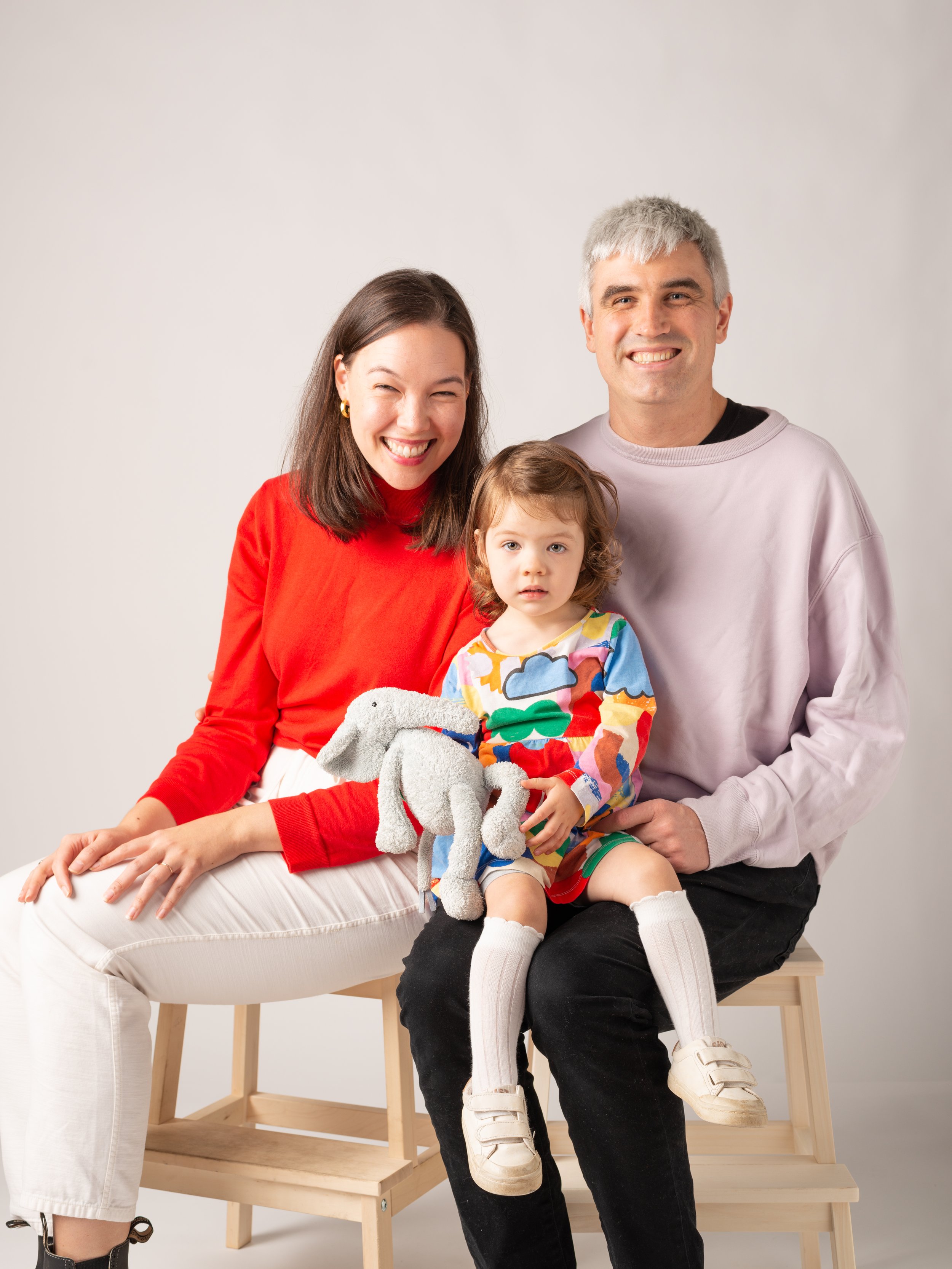 A smiling woman in a red sweater, a smiling man in a light gray sweatshirt, and a young girl with curly hair in a colorful dress sitting on a bench, holding a plush elephant toy, against a plain white background.