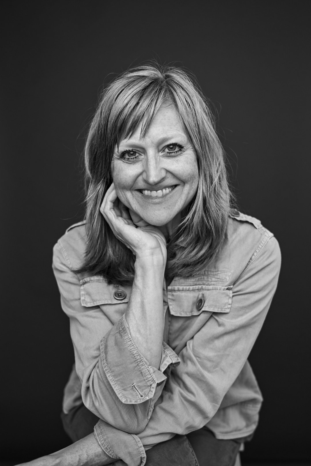 Black and white portrait of a smiling woman with shoulder-length hair, resting her chin on her hand, wearing a button-up shirt, against a dark background.