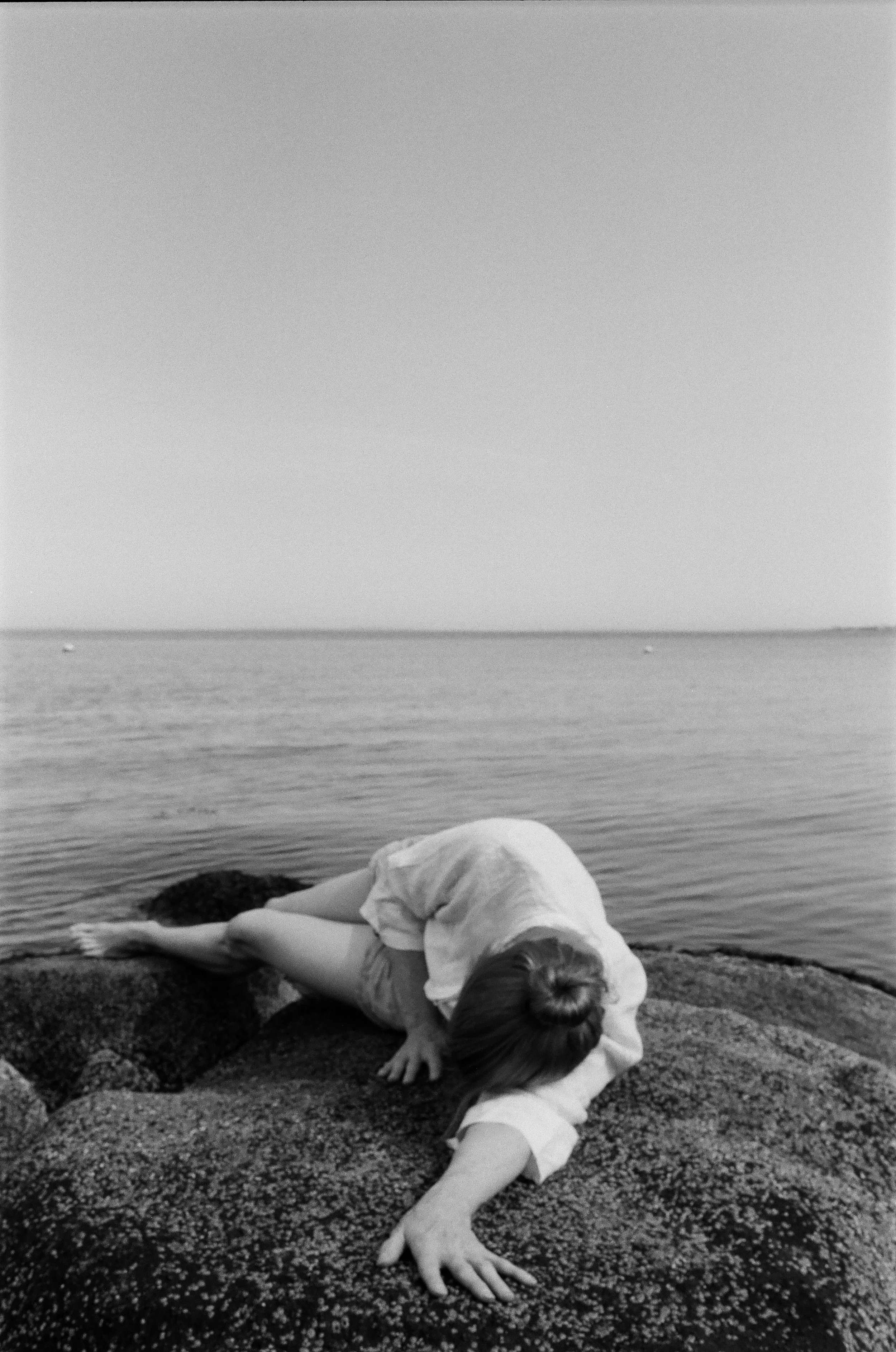 A person with long hair, lying on a large rock near the water, facing away from the camera, with their arm extended and hand resting on the rock.