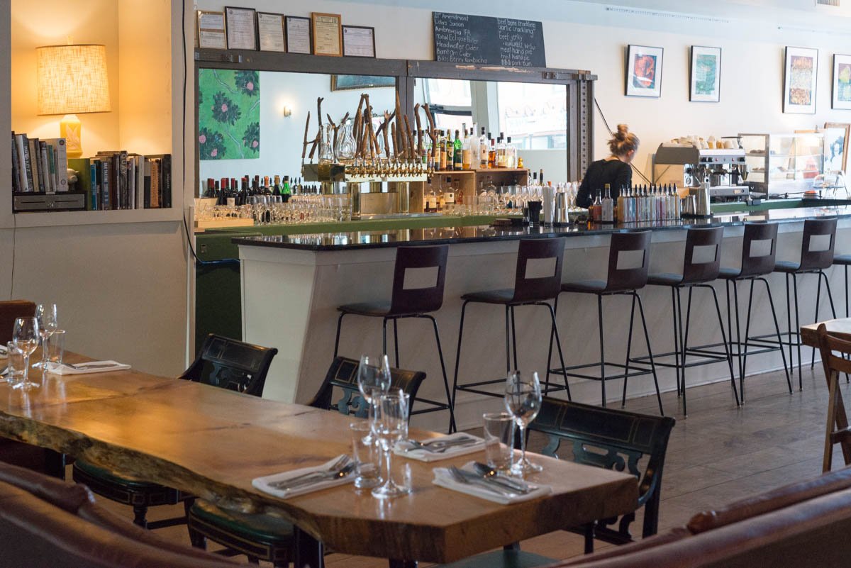 Interior of a restaurant bar with a row of barstools in front of the bar counter, glasses and bottles on the bar, a woman working behind the bar, and framed pictures on the wall.