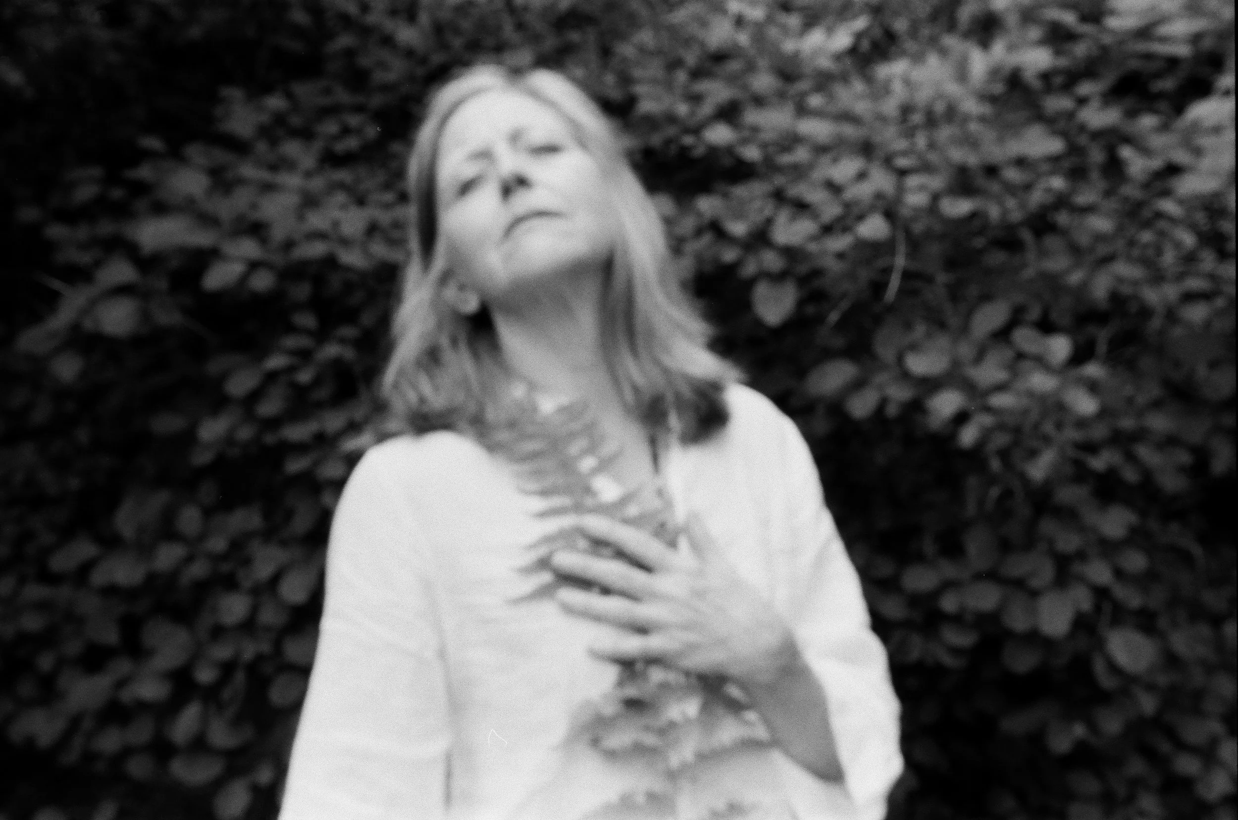 Black and white photo of a woman with her eyes closed, hand on her chest, standing in front of a background full of leaves.