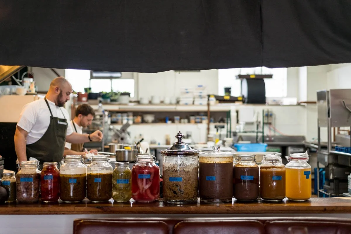 A restaurant kitchen with two chefs working behind a row of glass jars filled with various sauces and condiments on a wooden counter.