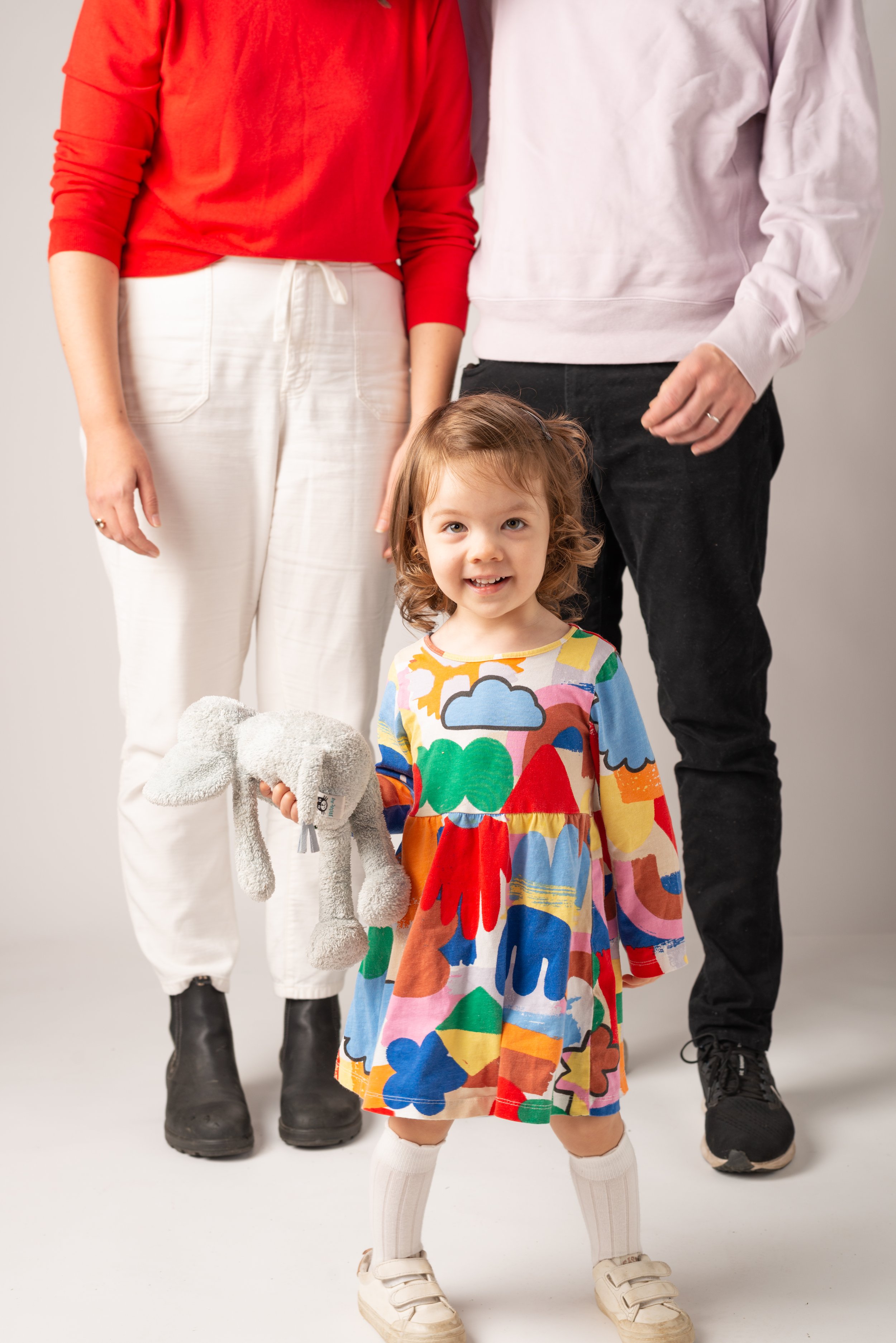 A young girl with curly hair standing in front of two adults, holding a plush toy elephant, wearing a colorful dress with abstract patterns and white knee-high socks.