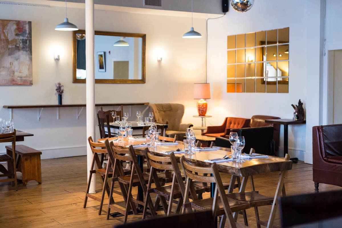 Interior of a restaurant with wooden tables and chairs, set with glasses and silverware, decorated with lamps, mirrors, and artwork on the walls.