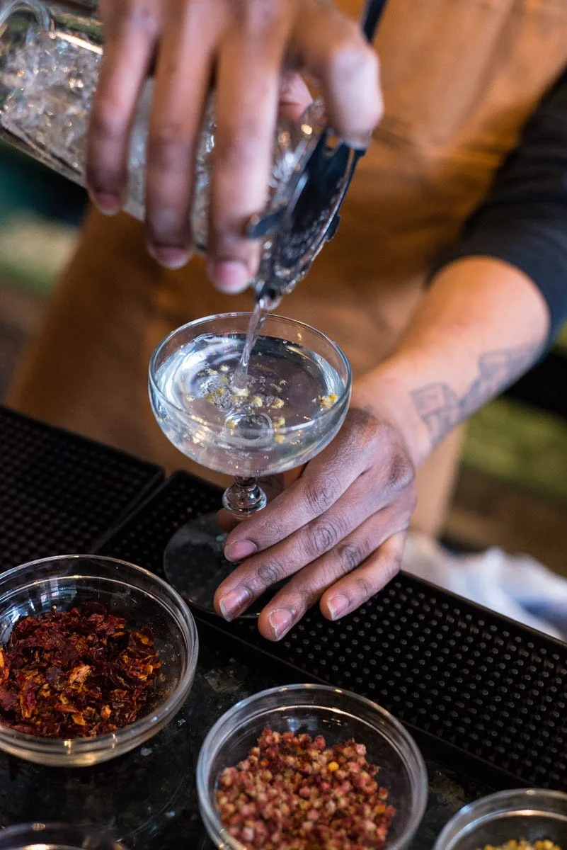 Person pouring clear liquid into a cocktail glass with yellow and purple flower petals. Multiple small bowls of colorful dried ingredients are on the table.