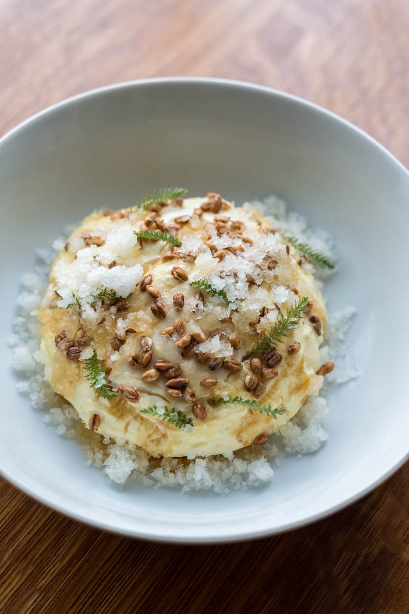 A bowl of ice cream topped with toasted seeds, crumbled white topping, and small green herbs, surrounded by crushed ice.