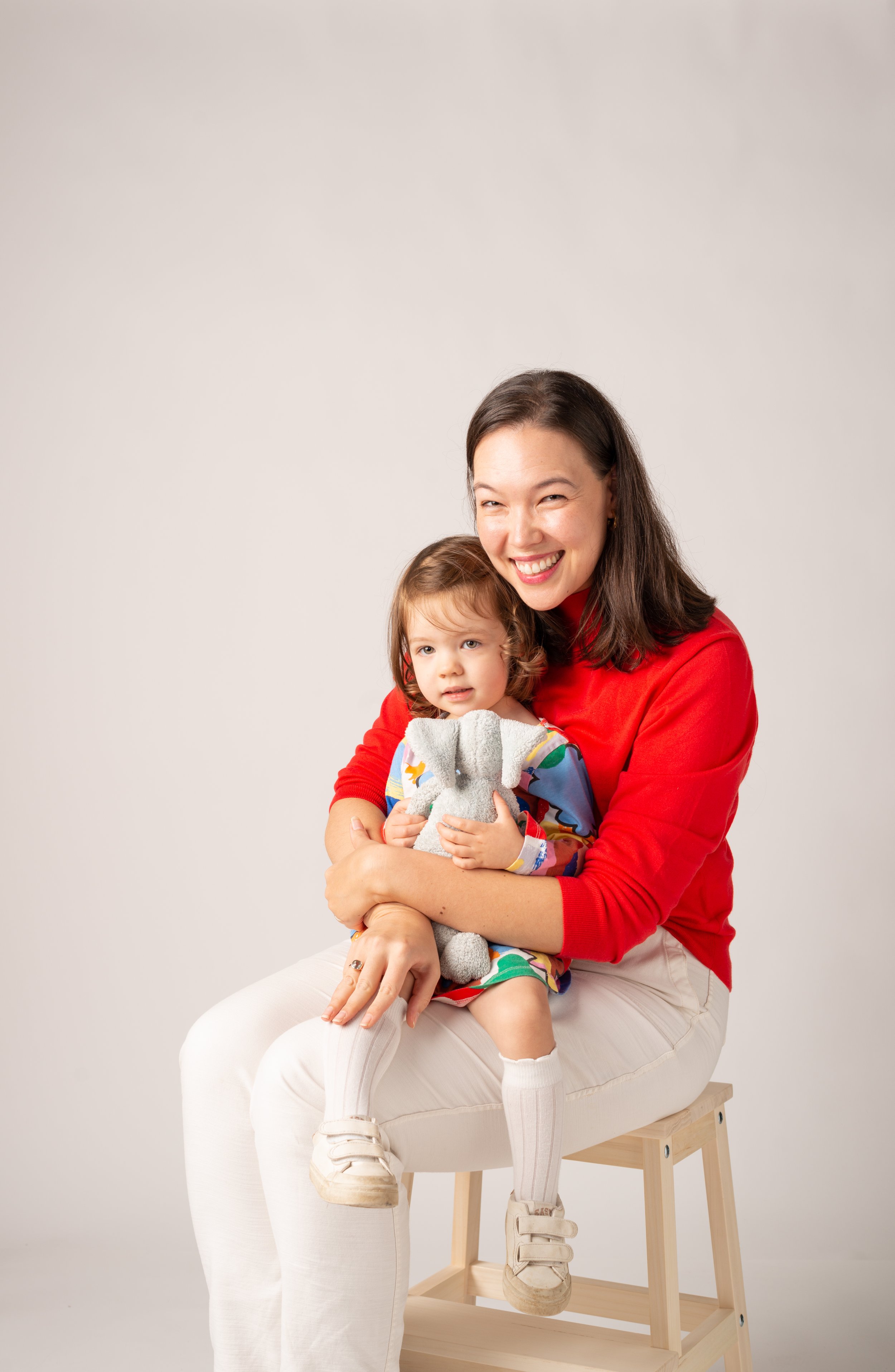 A young woman in a red sweater sitting on a stool, holding a young girl on her lap. The girl is holding a stuffed elephant toy. Both are smiling, with a plain light-colored background.