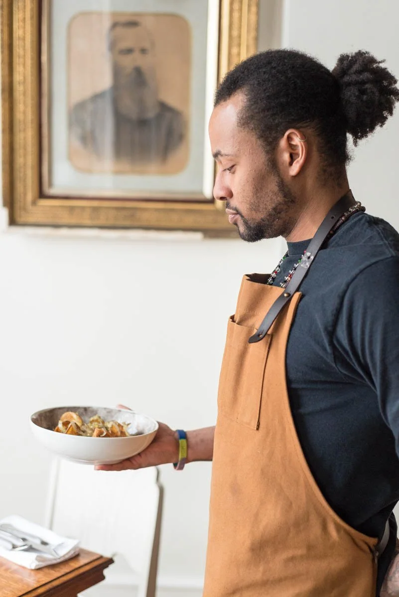 A man with dark curly hair tied back, wearing a black shirt and a tan apron, stands by a table holding a bowl of steaming food and looking at it thoughtfully. A framed black-and-white portrait is on the wall behind him.