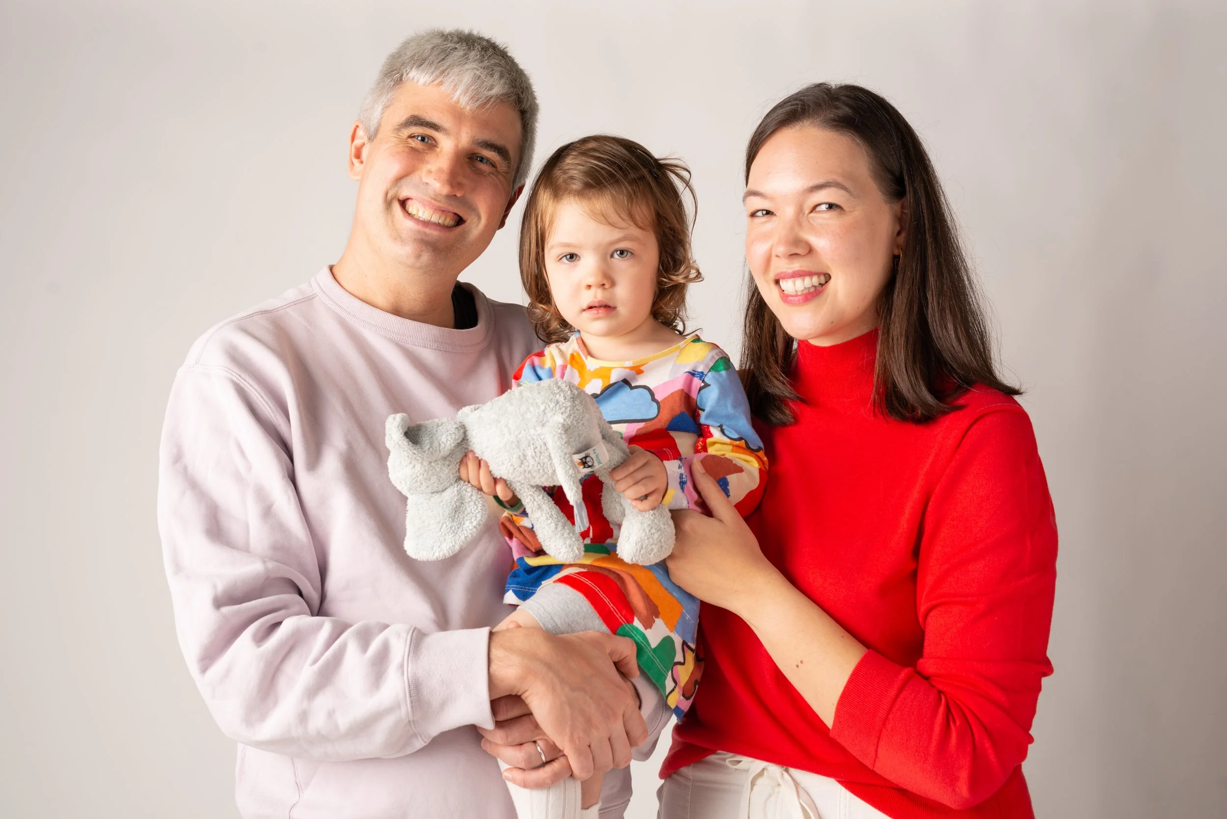 A family of three standing together, smiling, with a young girl holding a stuffed animal, against a plain light gray background.