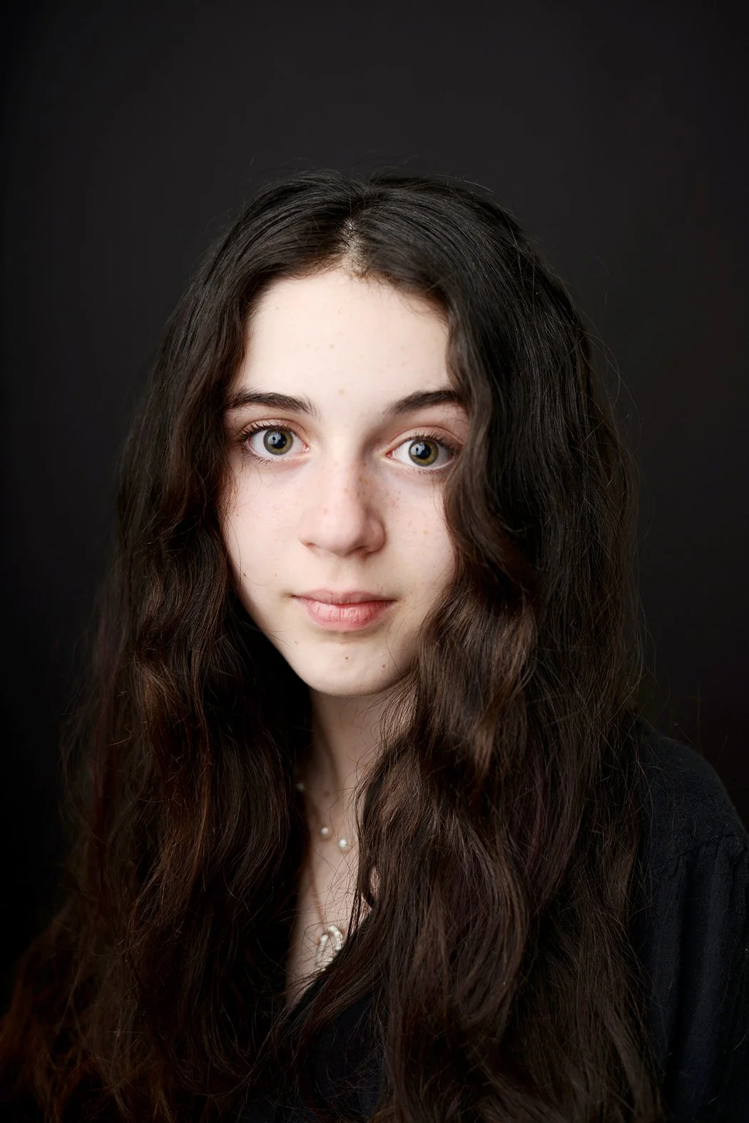 Portrait of a young woman with long, wavy brown hair and fair skin, against a dark background.