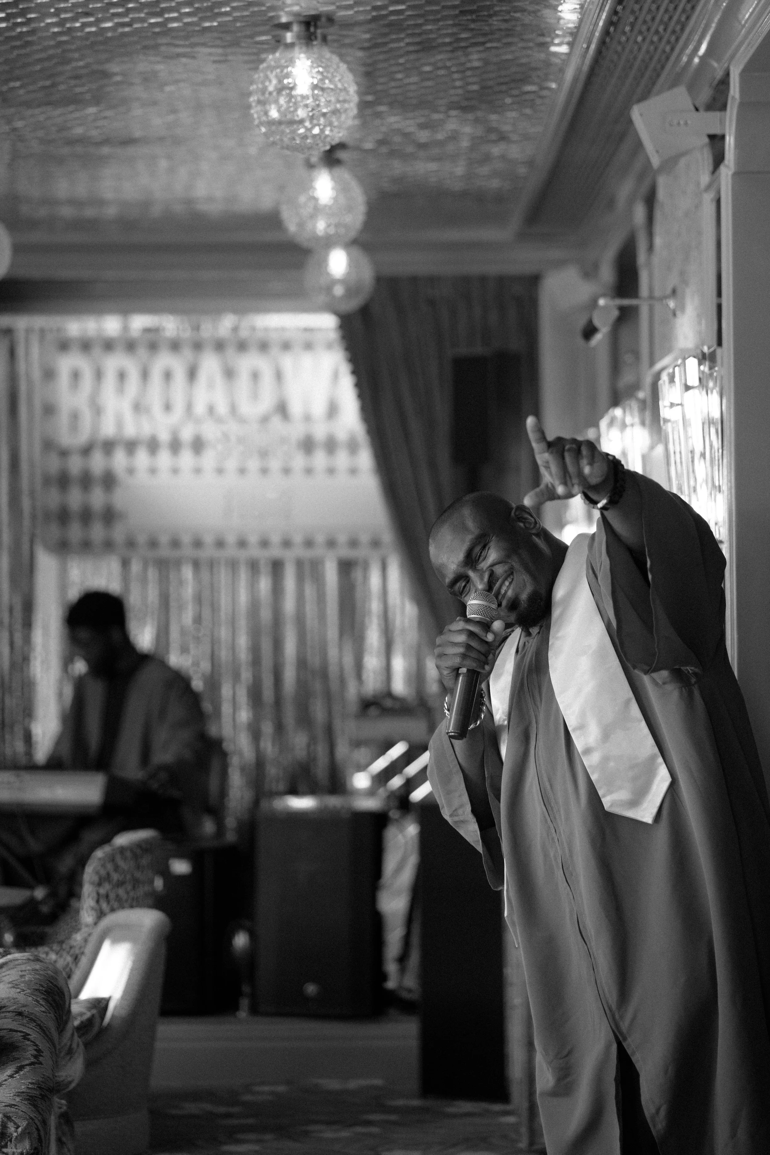 A man in a graduation gown singing into a microphone, with one hand raised, inside a decorated event hall.