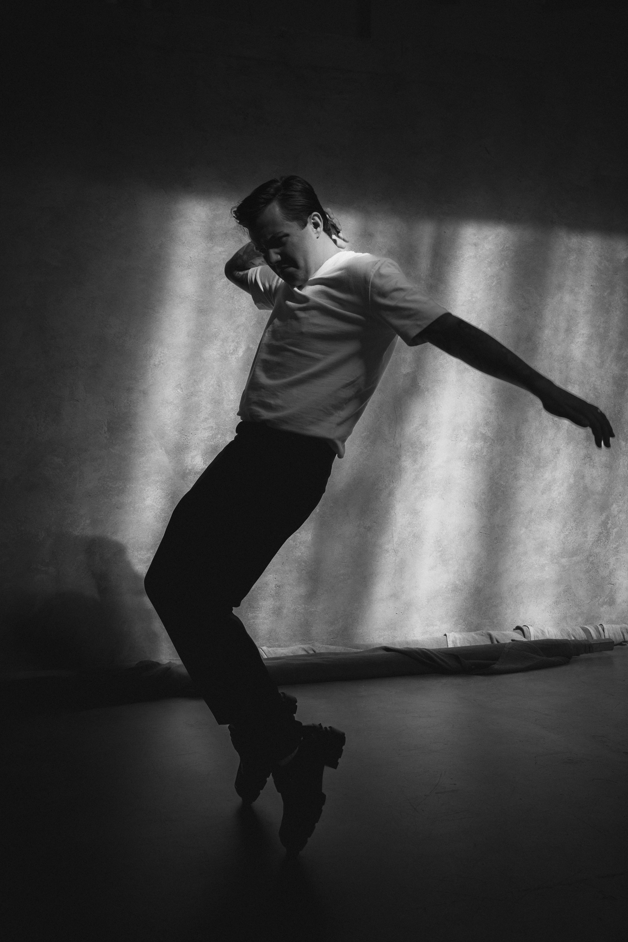 A black-and-white photo of a person roller skating indoors, balancing on one skate with arms extended, against a textured wall background.