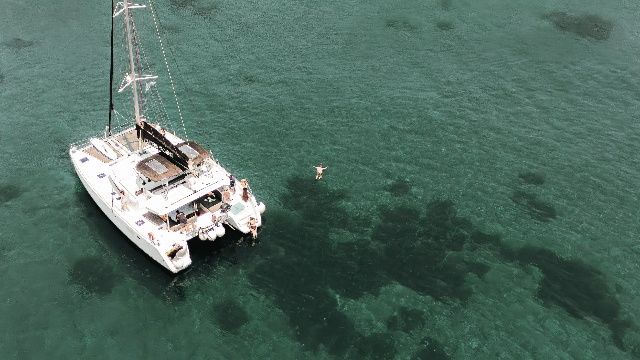 Bird's-eye view of a white yacht anchored in clear green water with people on board and one person swimming nearby.