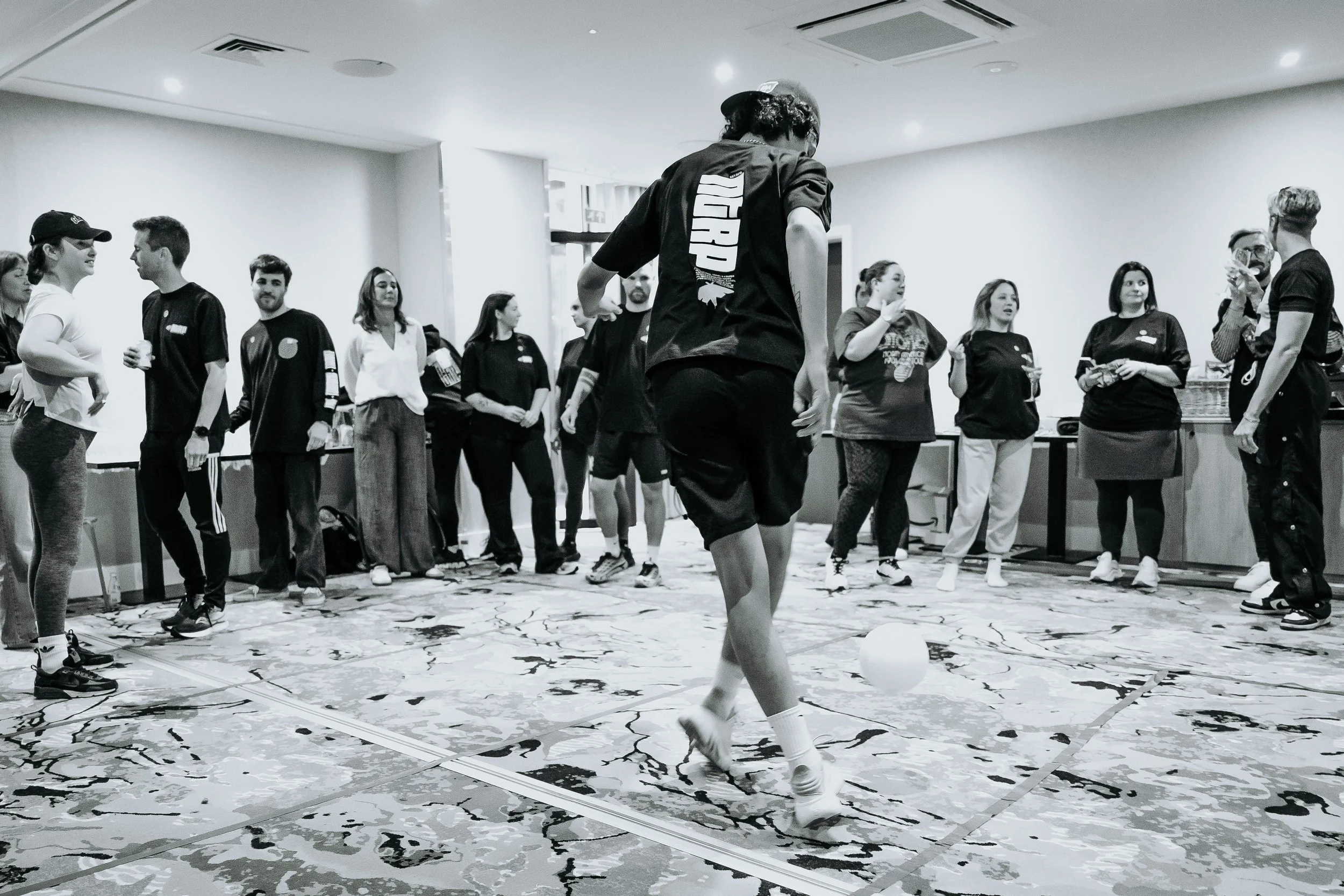 Group of people gathered in a room, casual attire, some holding drinks, one person barefoot playing with a ball on a patterned floor, others observing, ceiling lights, and air vents visible.