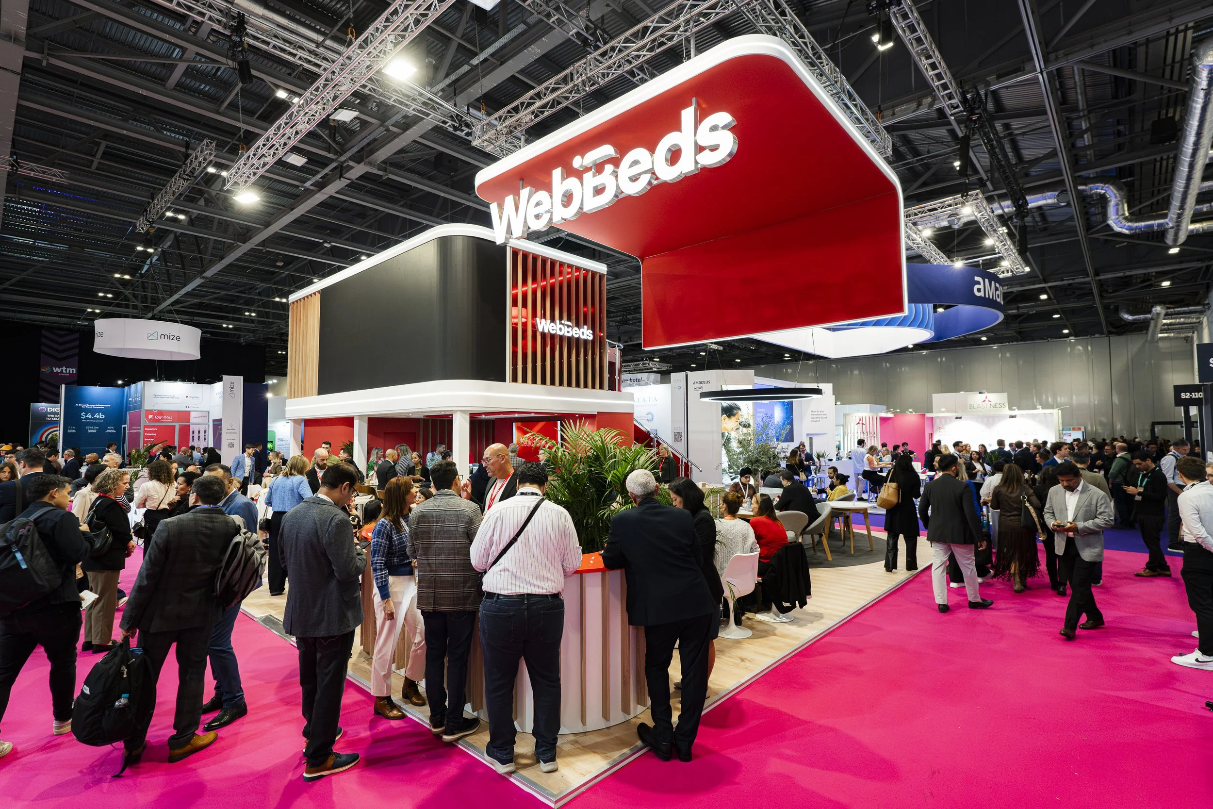 A crowded exhibition hall with a booth for Webbed, featuring a large red and white sign overhead and various seating areas, surrounded by many attendees exploring the event.