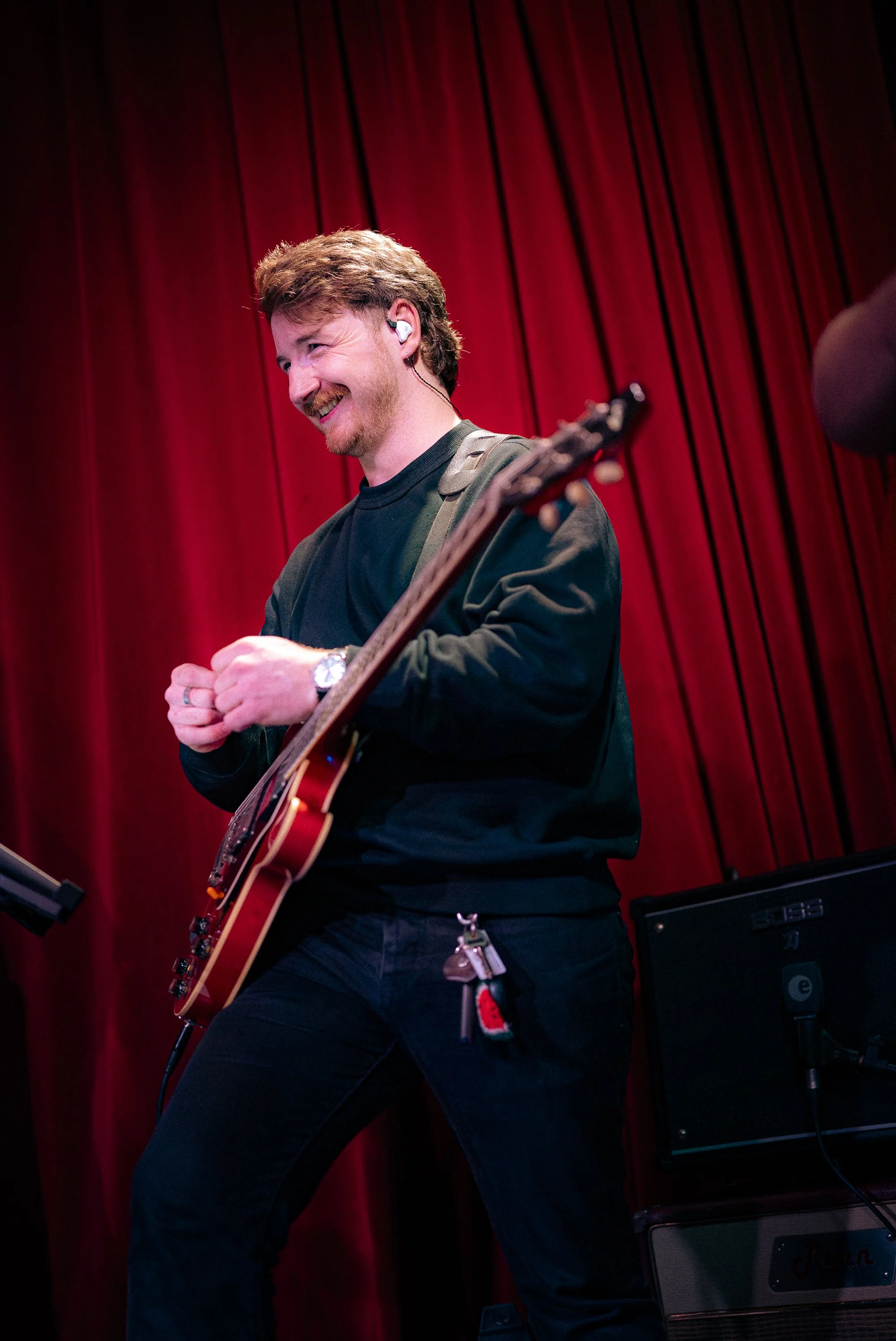 A man with red hair and a beard smiling while holding a guitar on stage with a red curtain background.