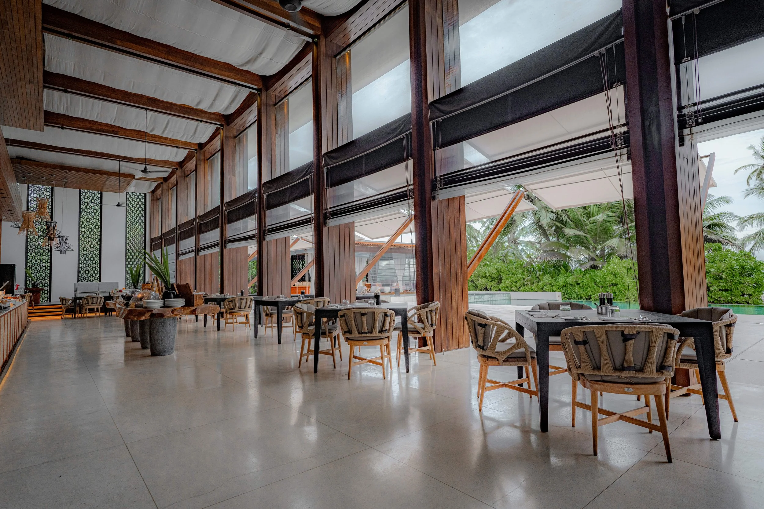 Empty restaurant with large windows overlooking a tropical garden, wooden and black furniture, and white ceiling with hanging lights.