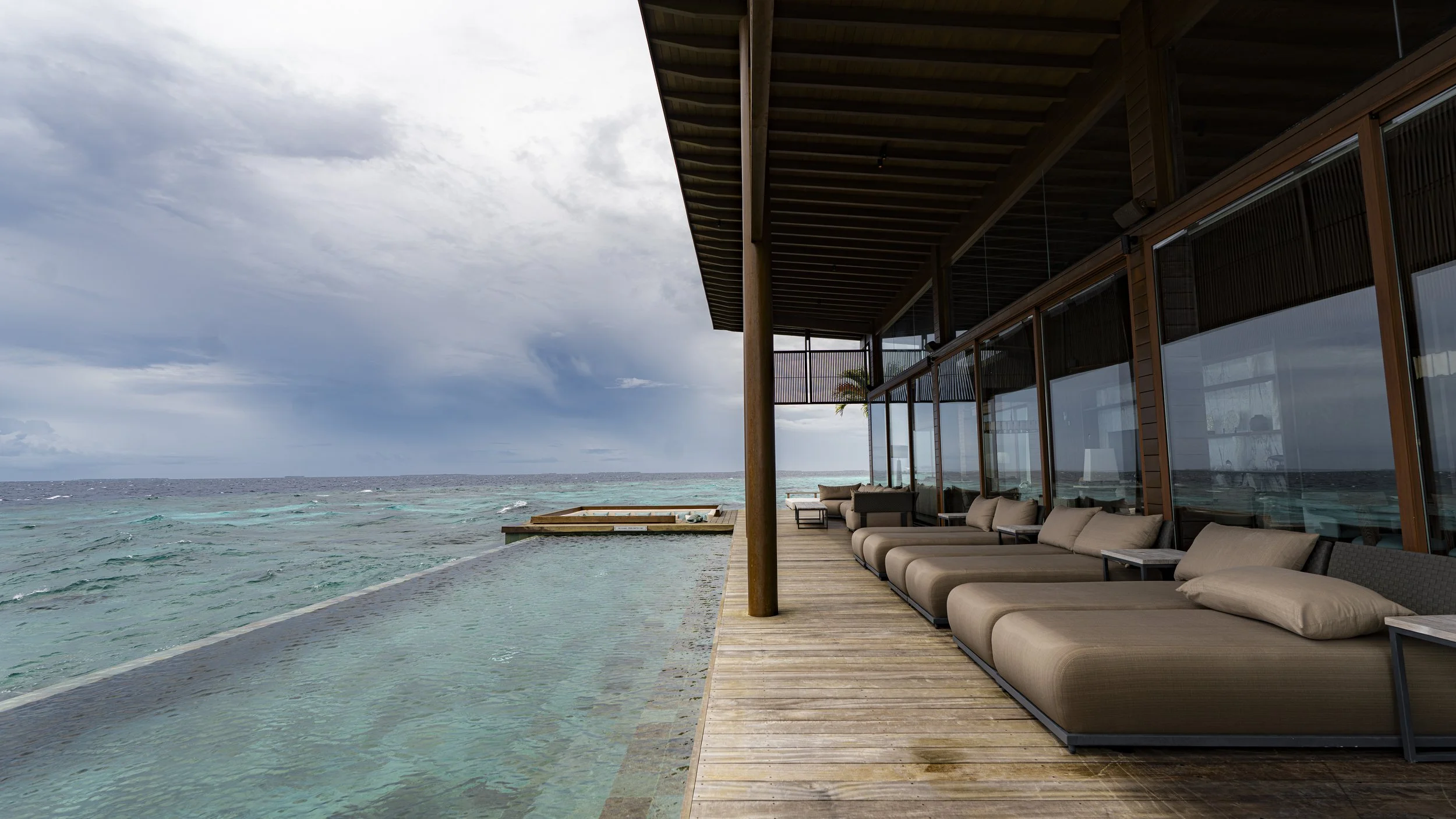 Outdoor lounge area with cushioned loungers on a wooden deck, overlooking the ocean with cloudy sky.