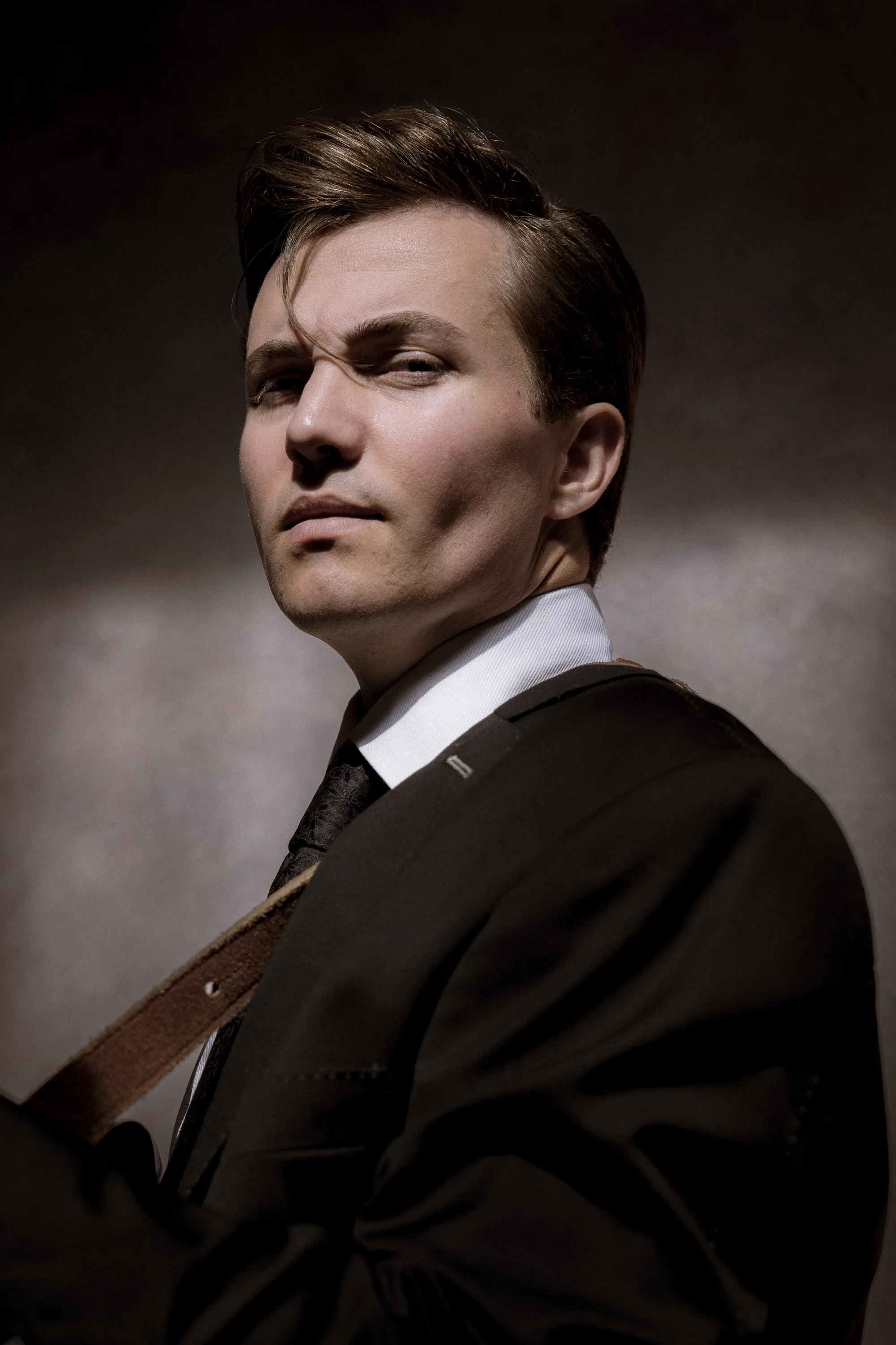 Close-up portrait of a young man with styled hair, wearing a formal black suit and white shirt, posing with a confident expression.