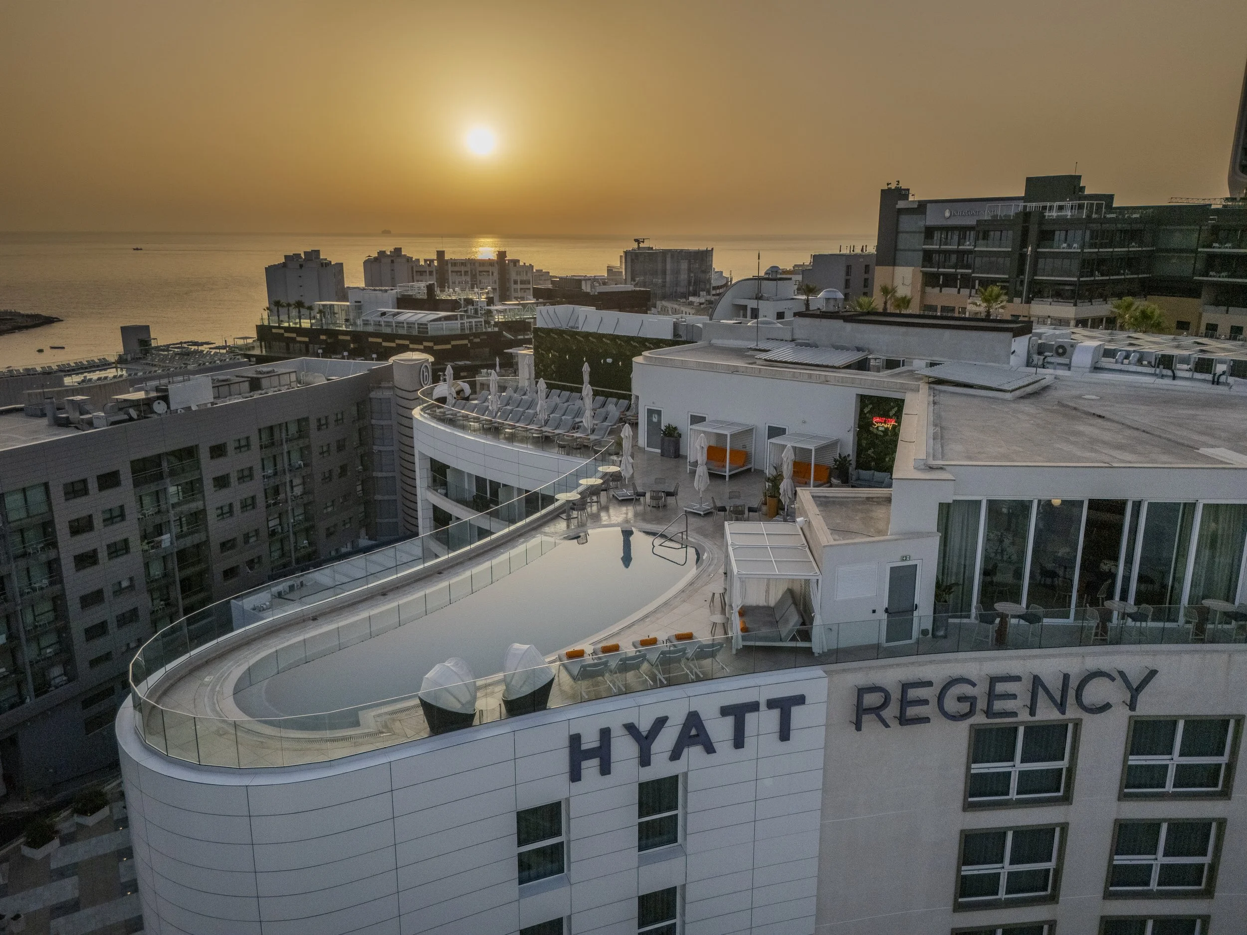 Sunset view over the rooftop pool and outdoor seating area at the Hyatt Regency hotel, with buildings and ocean in the background.