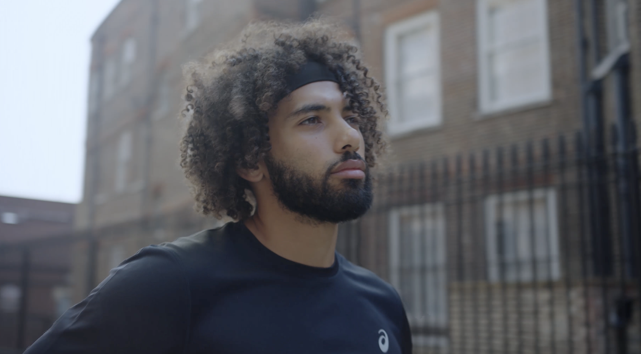 A man with curly hair and a beard wearing a black shirt and a headband, standing outdoors in an urban area with brick buildings and a metal fence in the background.