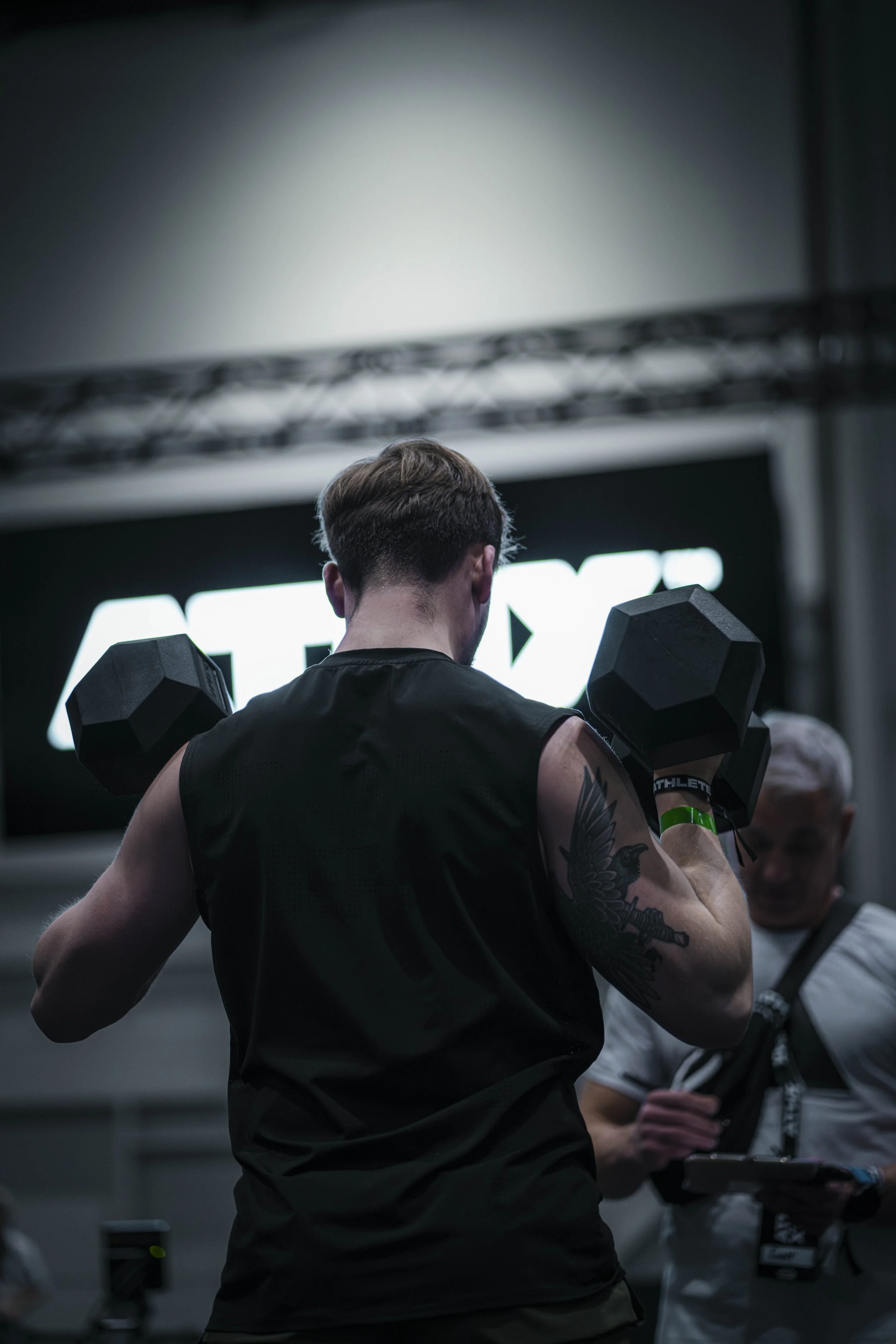 A man lifts dumbbells at a gym, seen from behind, with a large illuminated sign in the background.