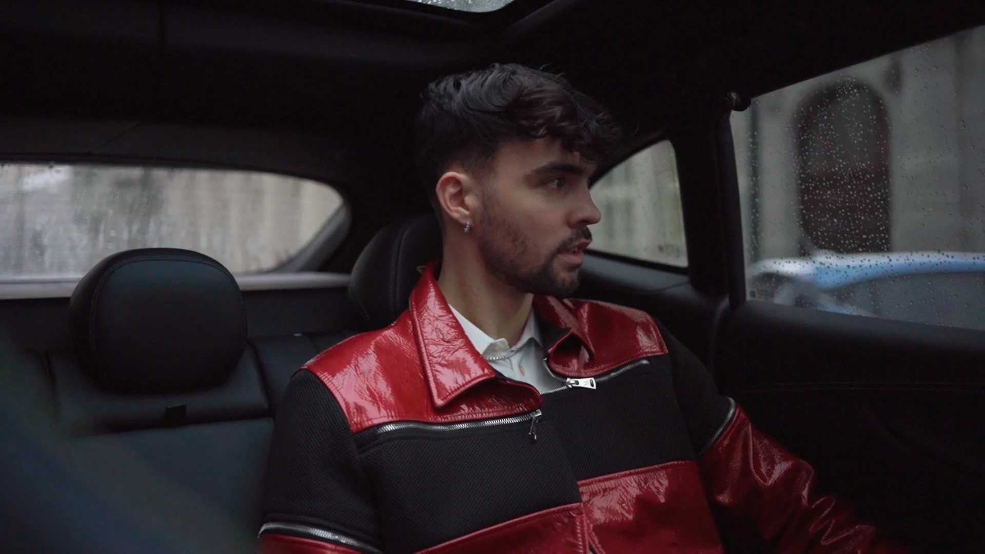 A young man with dark hair, beard, and earrings, wearing a red and black jacket, sitting in the backseat of a car during rainy weather.