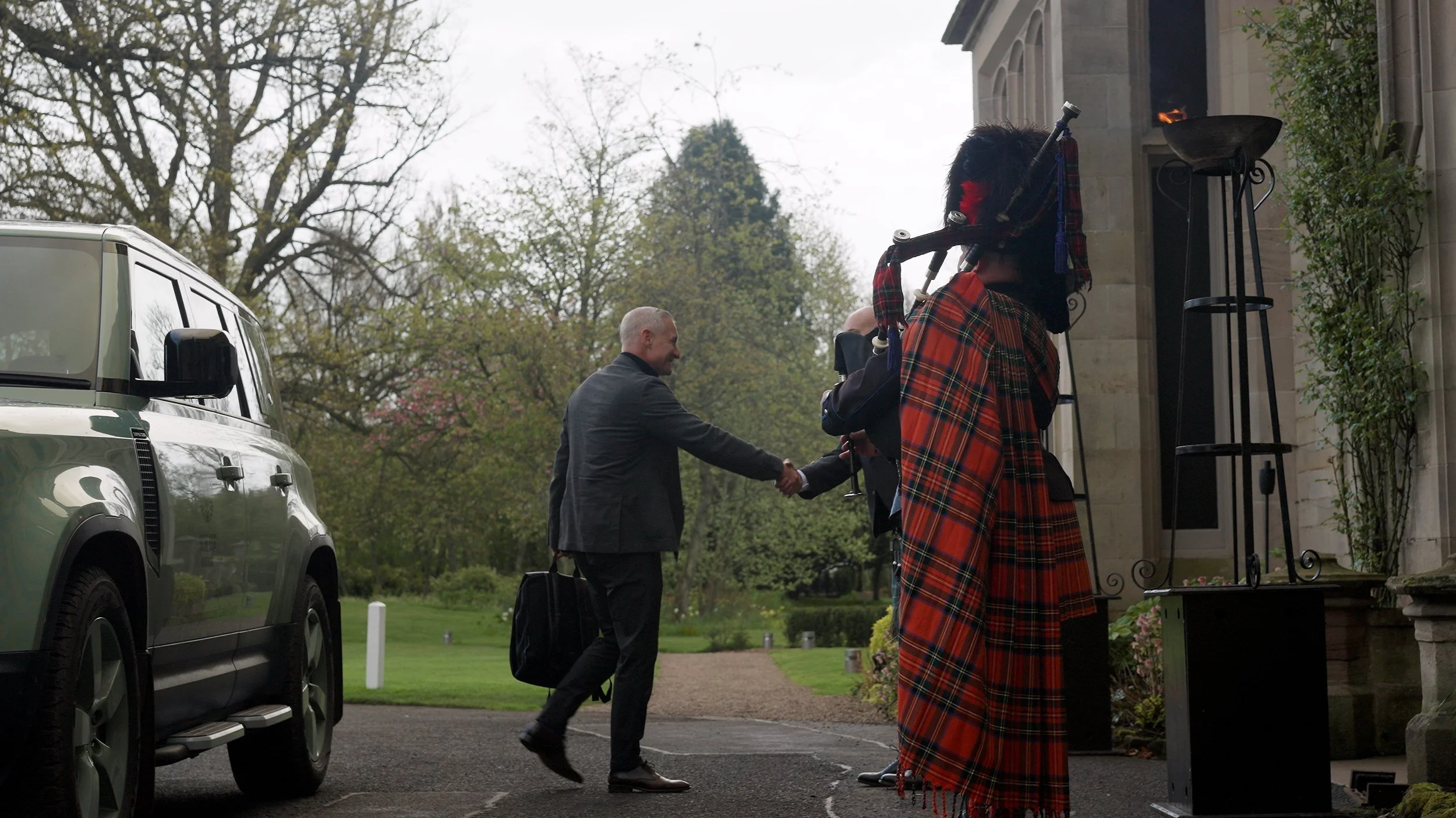 Two men shaking hands outside a building, one carrying a black bag; a man in traditional Scottish attire playing bagpipes nearby; a white vehicle parked on the left; trees and garden in the background.