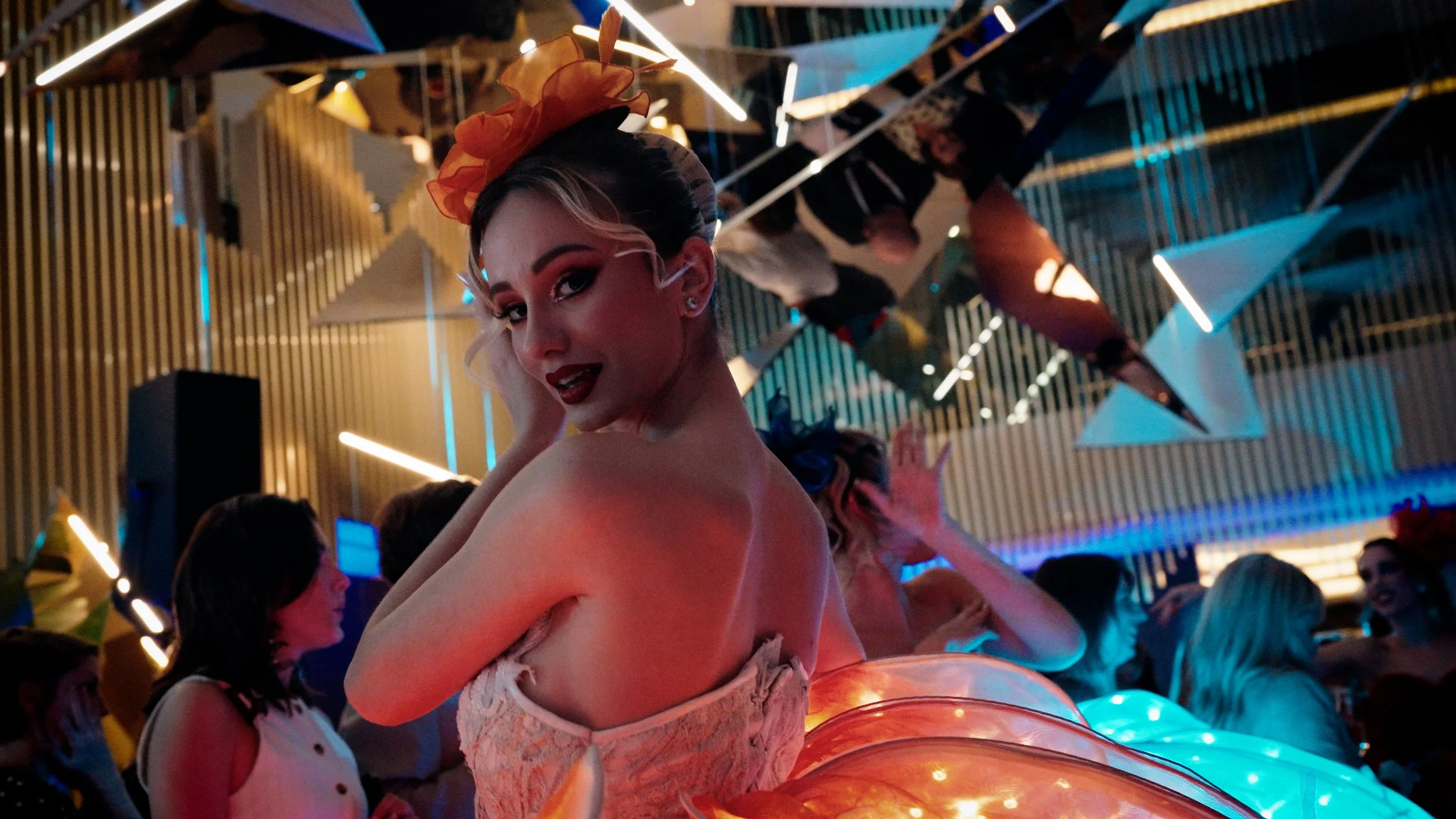 A woman with styled hair and makeup wearing a strapless dress and a large yellow bow headpiece, posing with her hand on her face in a lively, colorful event or party with abstract hanging decorations and ambient lighting.