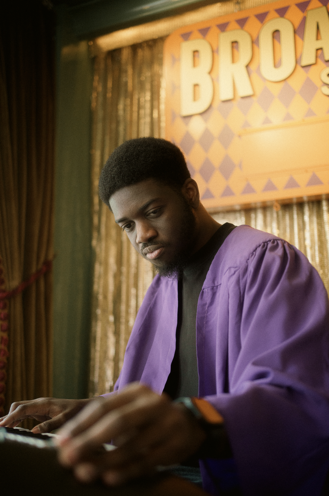 A young man in a purple graduation gown playing a keyboard at a graduation event, with a large orange and purple sign behind him that reads 'BROAD' and gold curtains in the background.