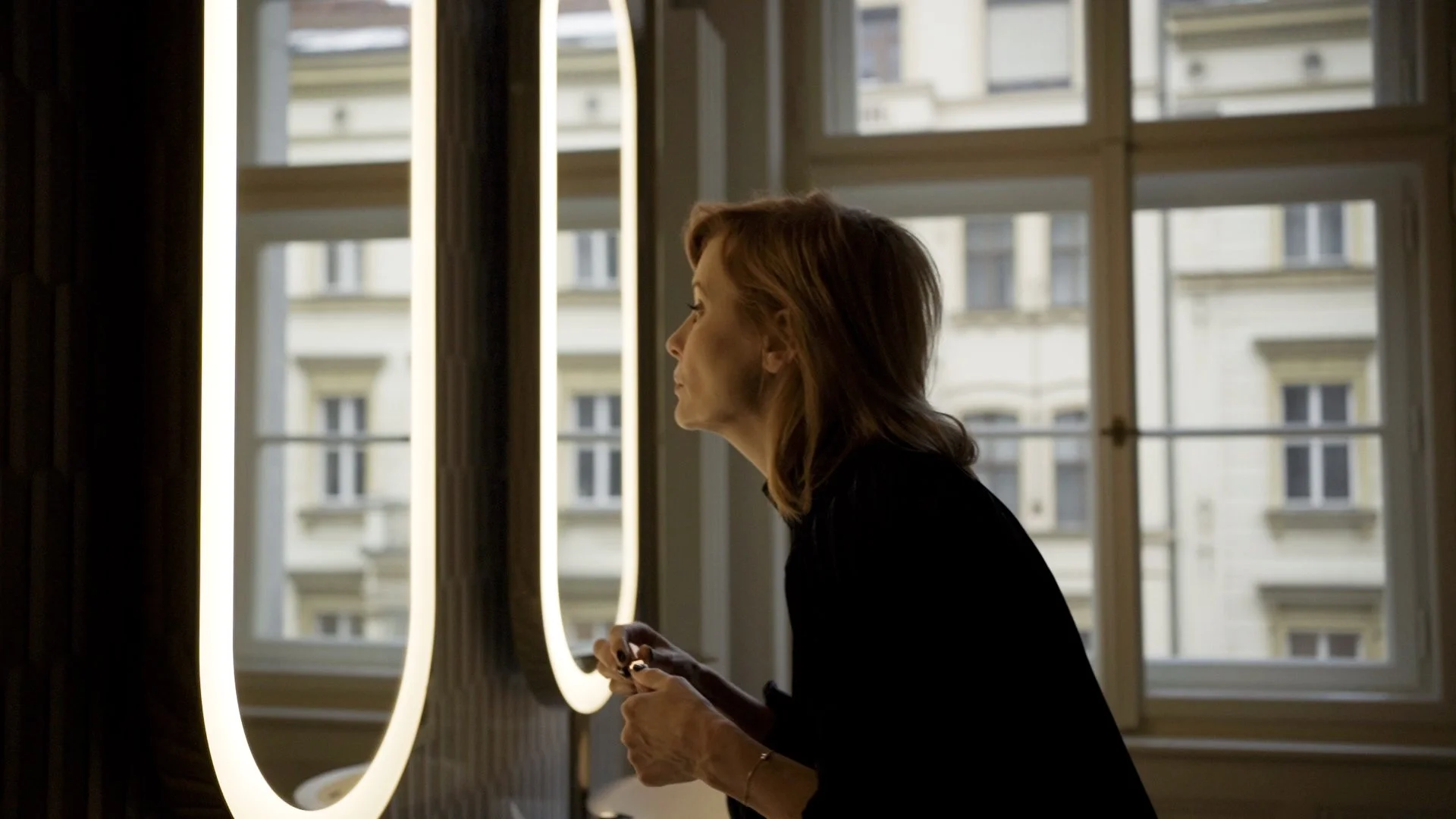 A woman with shoulder-length hair is standing near a window, holding a phone, with the city buildings visible outside.