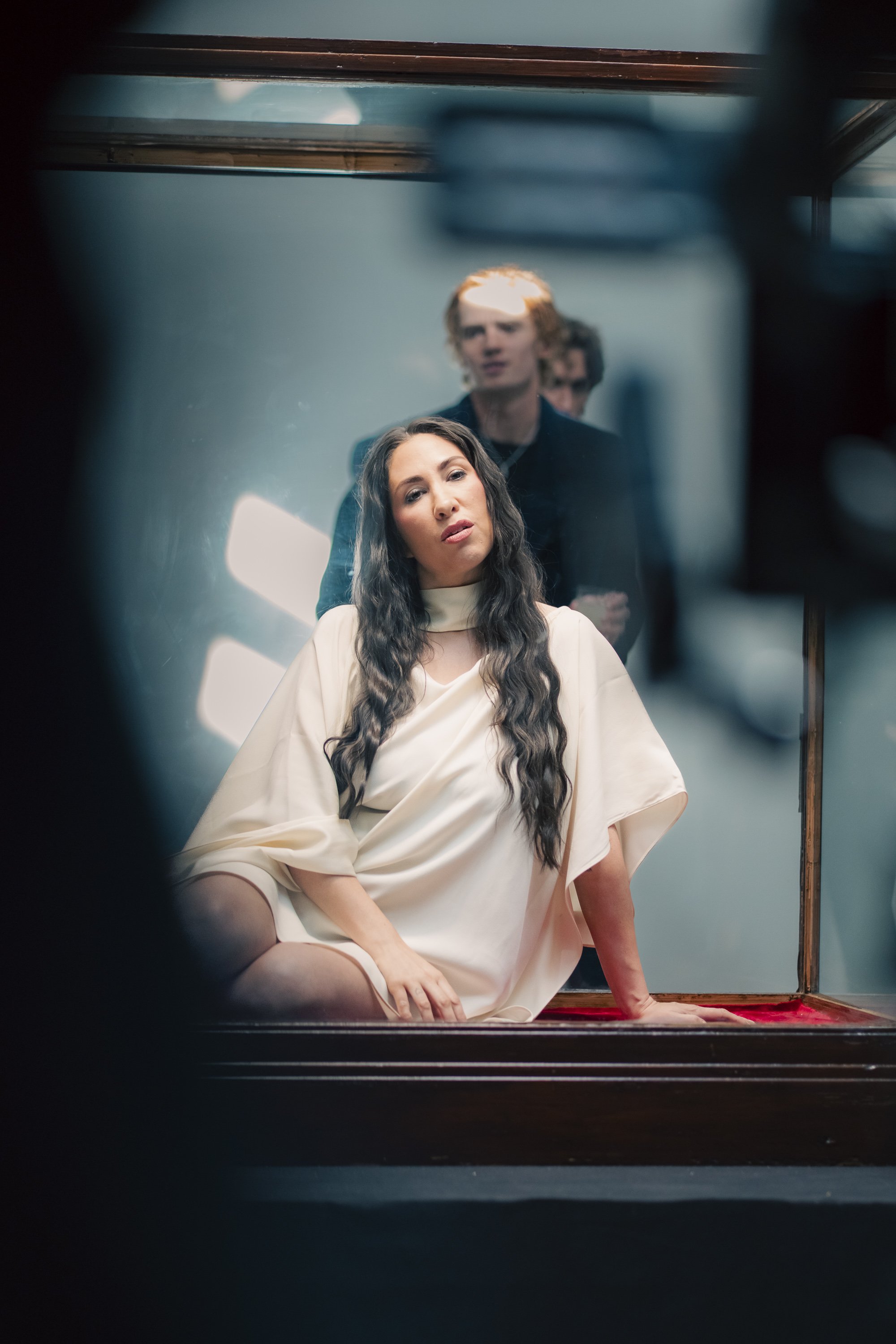 A woman with long dark wavy hair and a white dress poses on a vintage red velvet settee, as seen through a mirror with a blurred reflection of two men standing behind her.