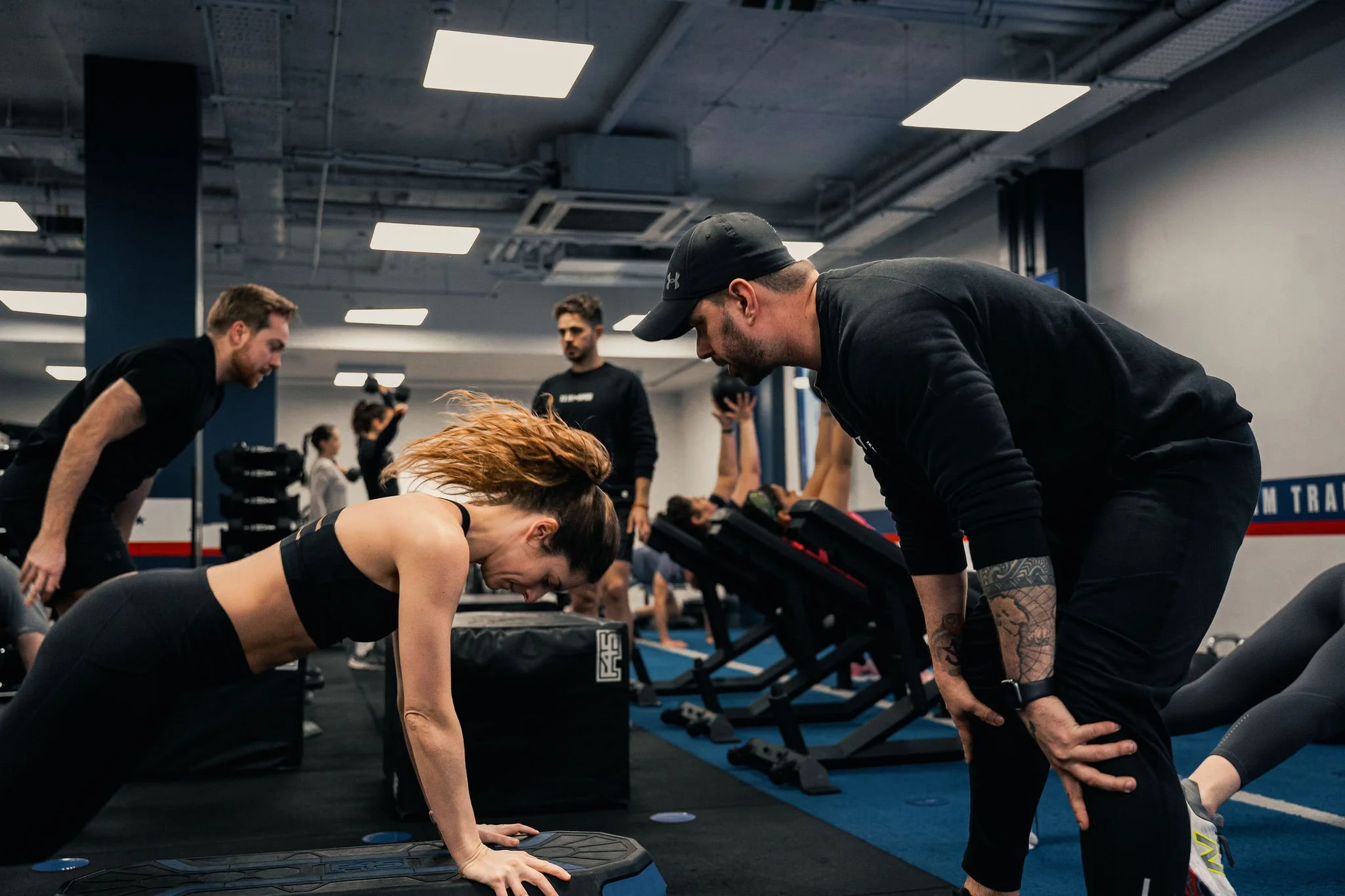 A group fitness class in a gym with a woman doing push-ups, instructor guiding her, other participants exercising in the background.