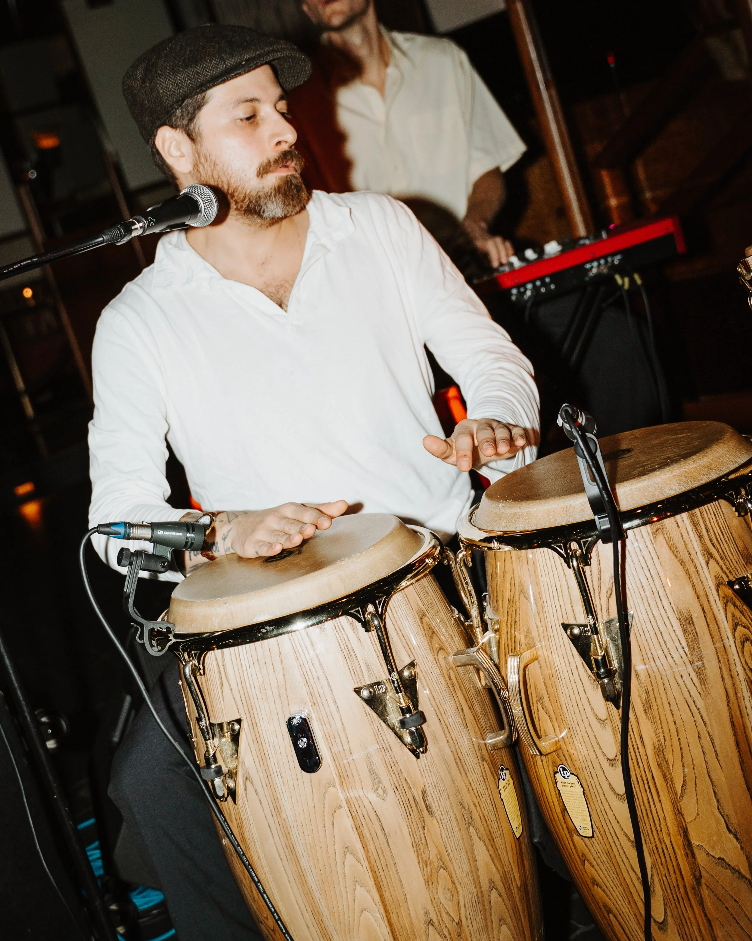 A man playing bongos, wearing a white shirt and a flat cap, with a beard and mustache, in a dimly lit setting with a person in the background playing a keyboard.