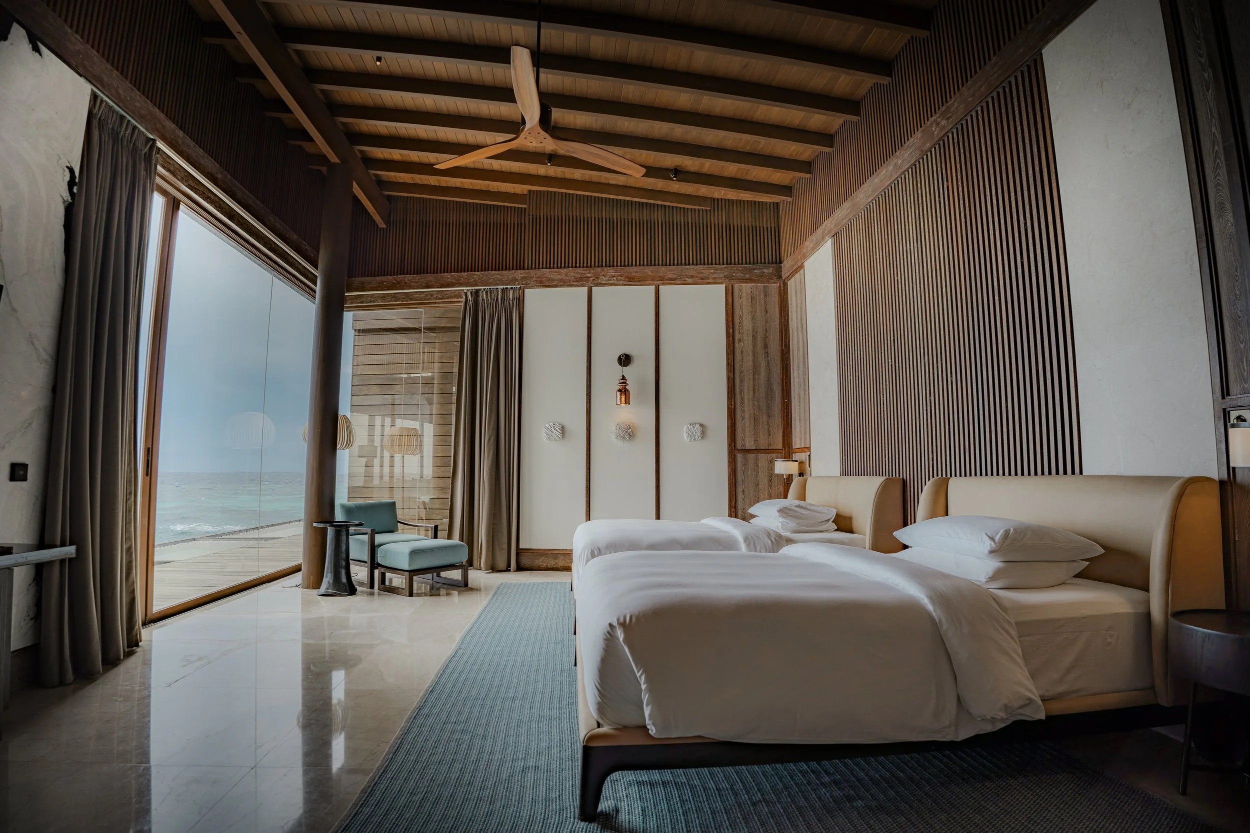 A modern beachside hotel room with two single beds, large sliding glass doors opening to an ocean view, wooden ceiling, and stylish decor.