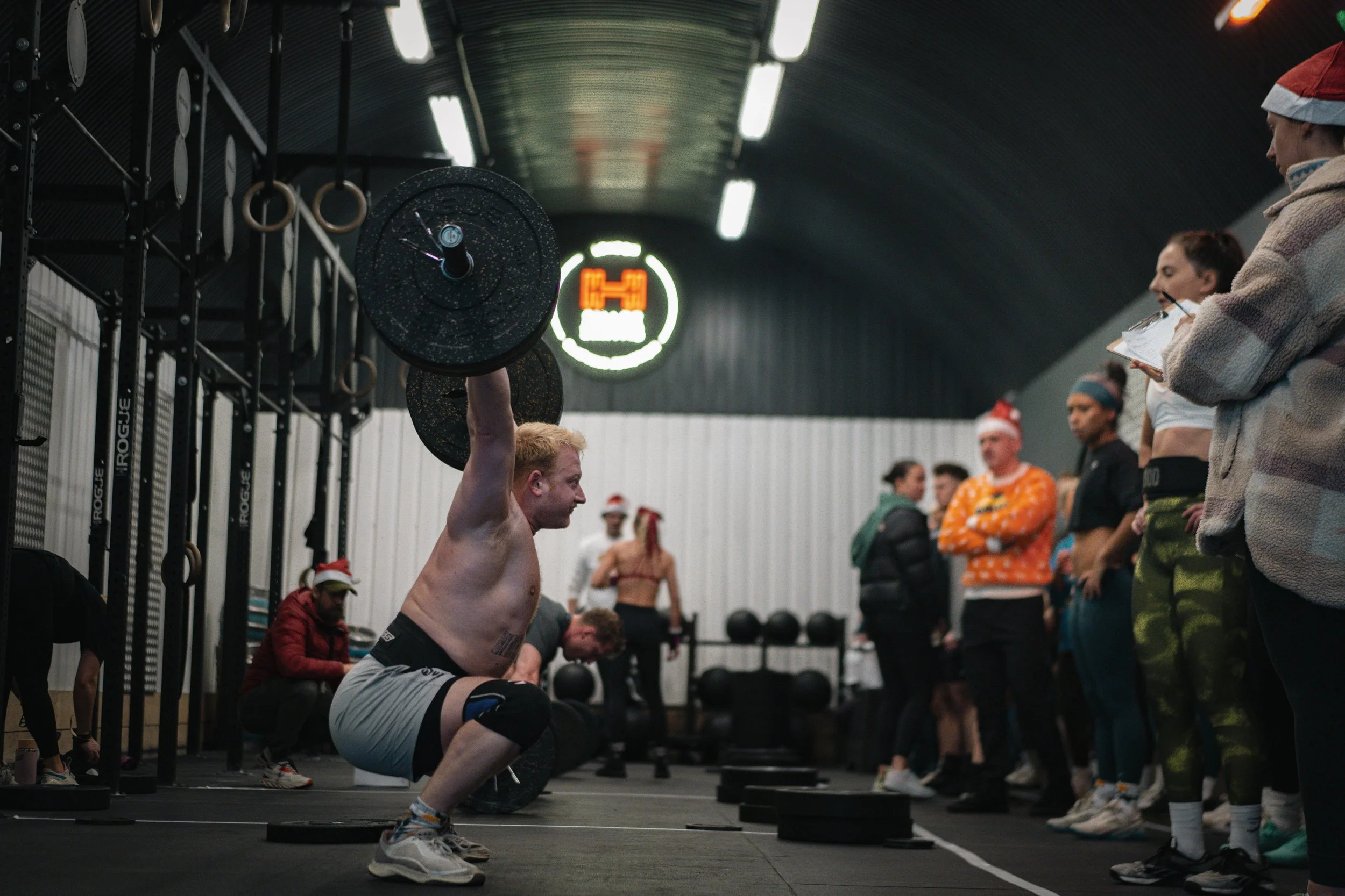 A man is performing a snatch lift during a CrossFit workout in a gym, surrounded by people watching and taking notes.