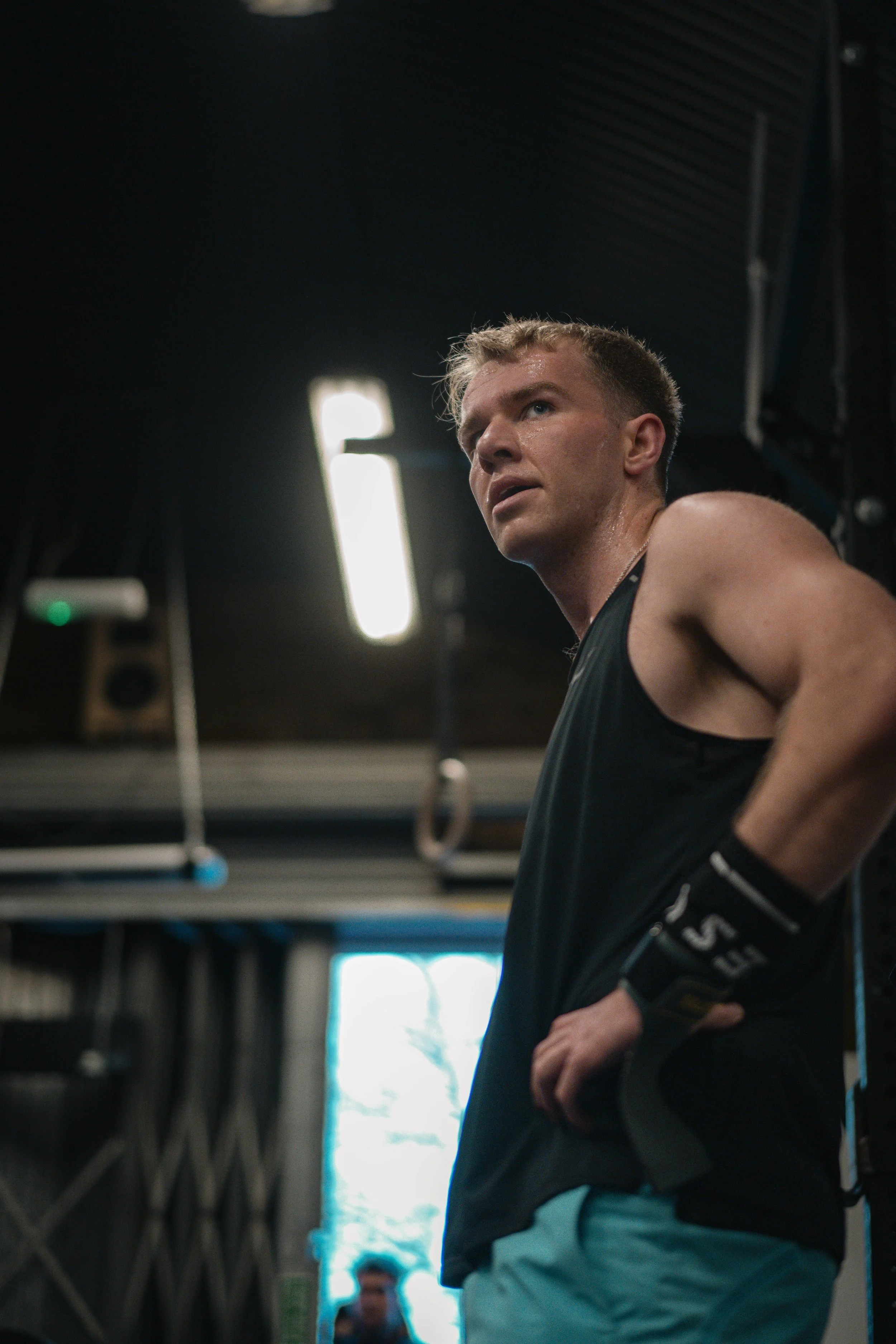 An athletic man in a gym, wearing a black tank top and workout gloves, appears tired and sweaty after exercising.