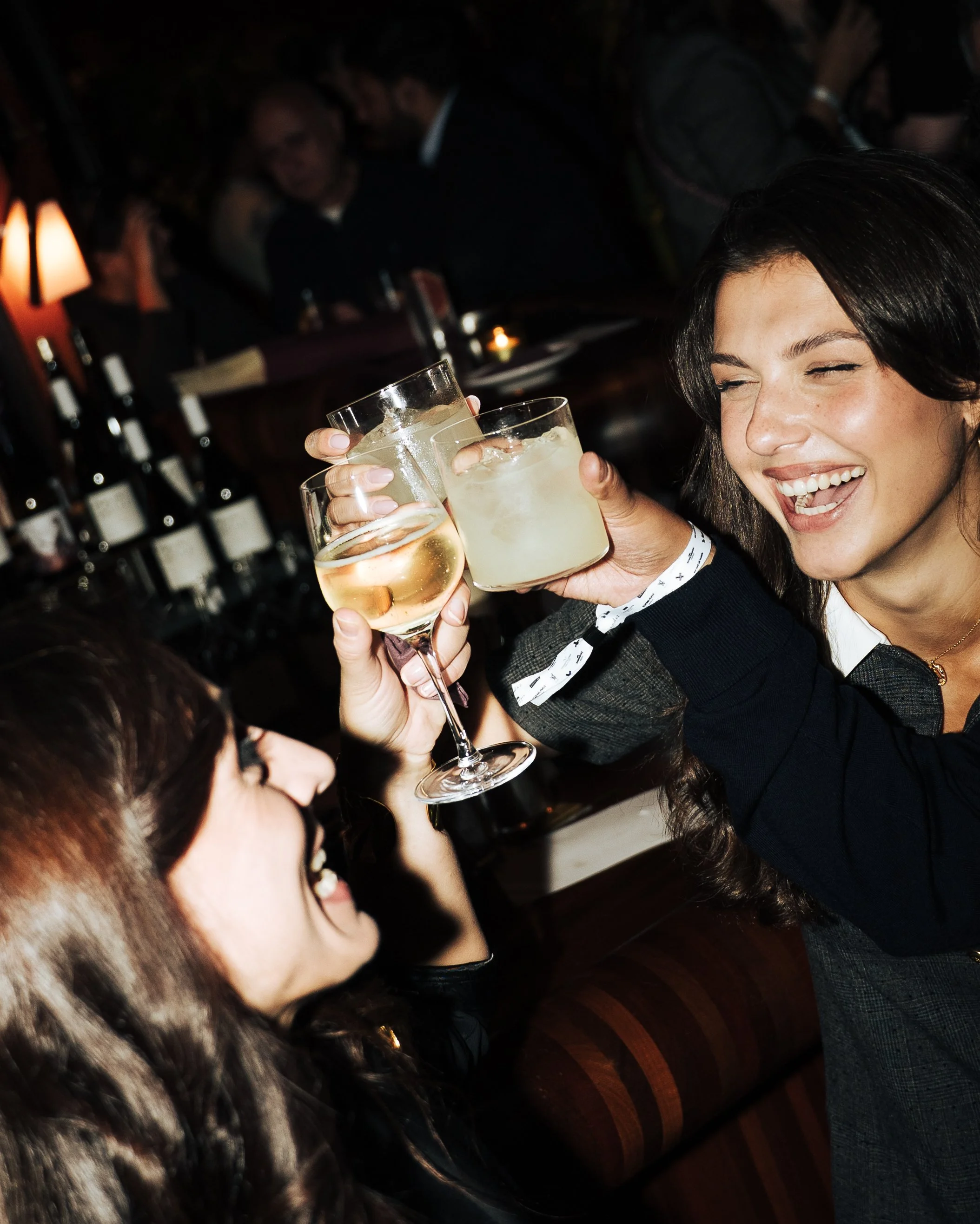 Two women clinking glasses of wine and a cocktail in a bar or restaurant, smiling and enjoying themselves.