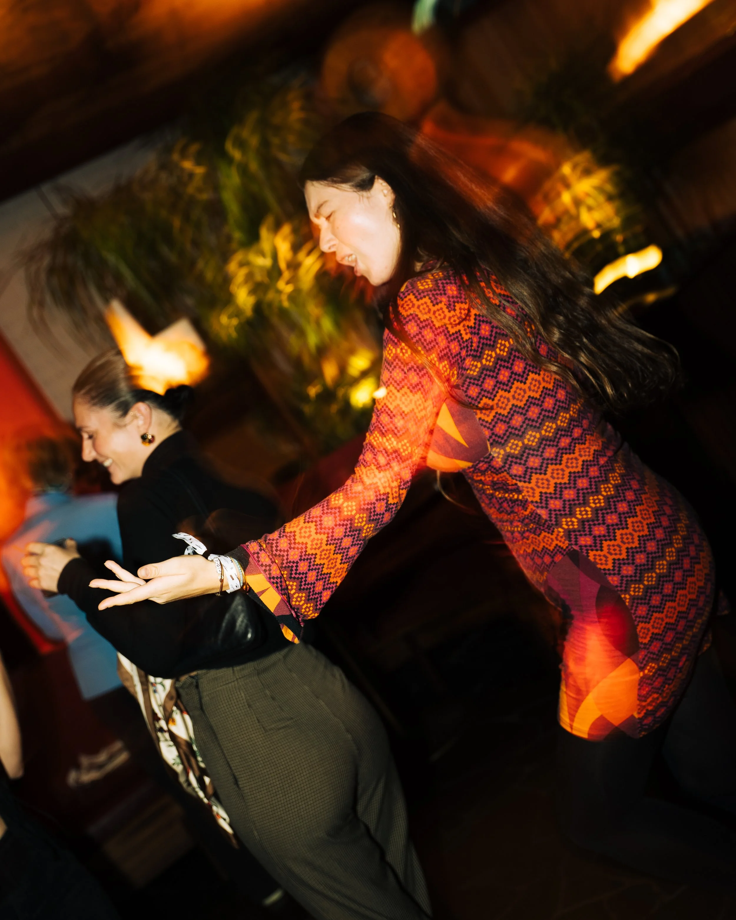 Two women dancing happily in a lively, dimly lit environment with colorful lights and blurred background.