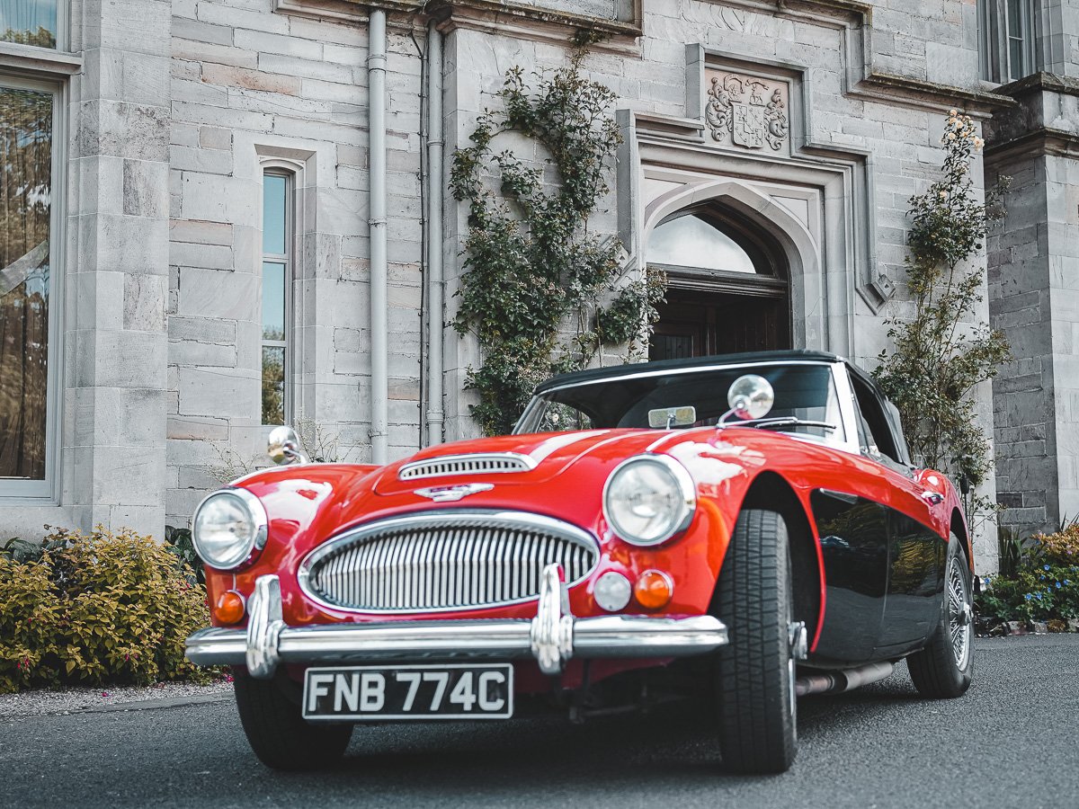 A classic red and black vintage car parked in front of a stone building with vines and plants around the entrance.